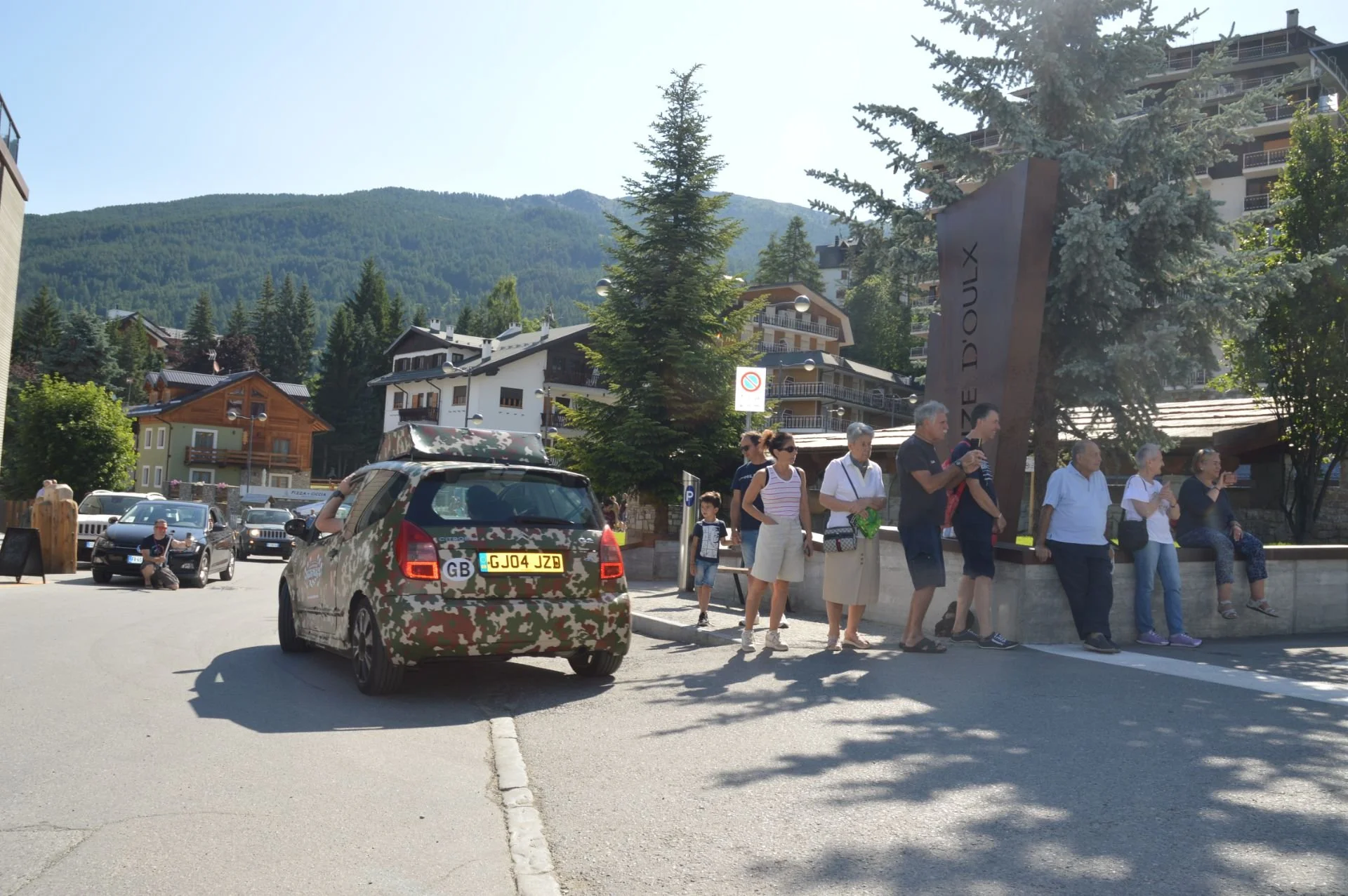 People waiting in line near a large monument in a mountain town with houses and trees, a car in camouflage pattern in the foreground, under sunny weather.