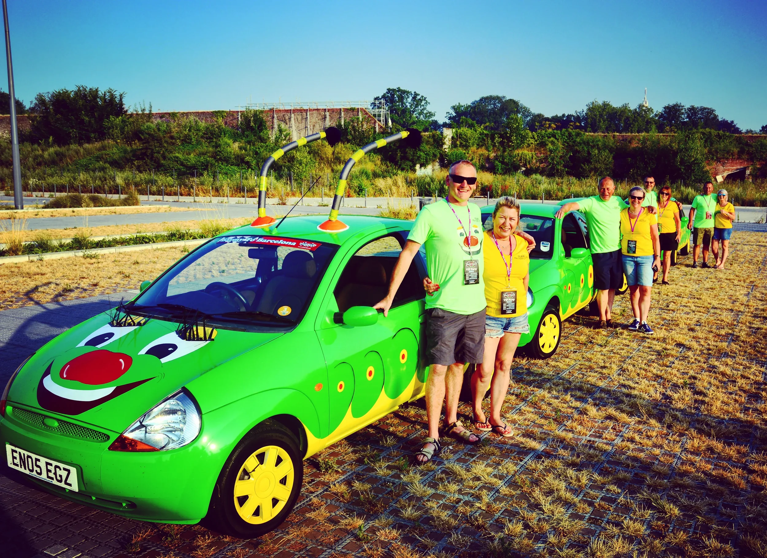 Group of people standing in a line outside next to a bright green cartoon-themed car with a friendly face on the hood, on a sunny day with clear blue sky.
