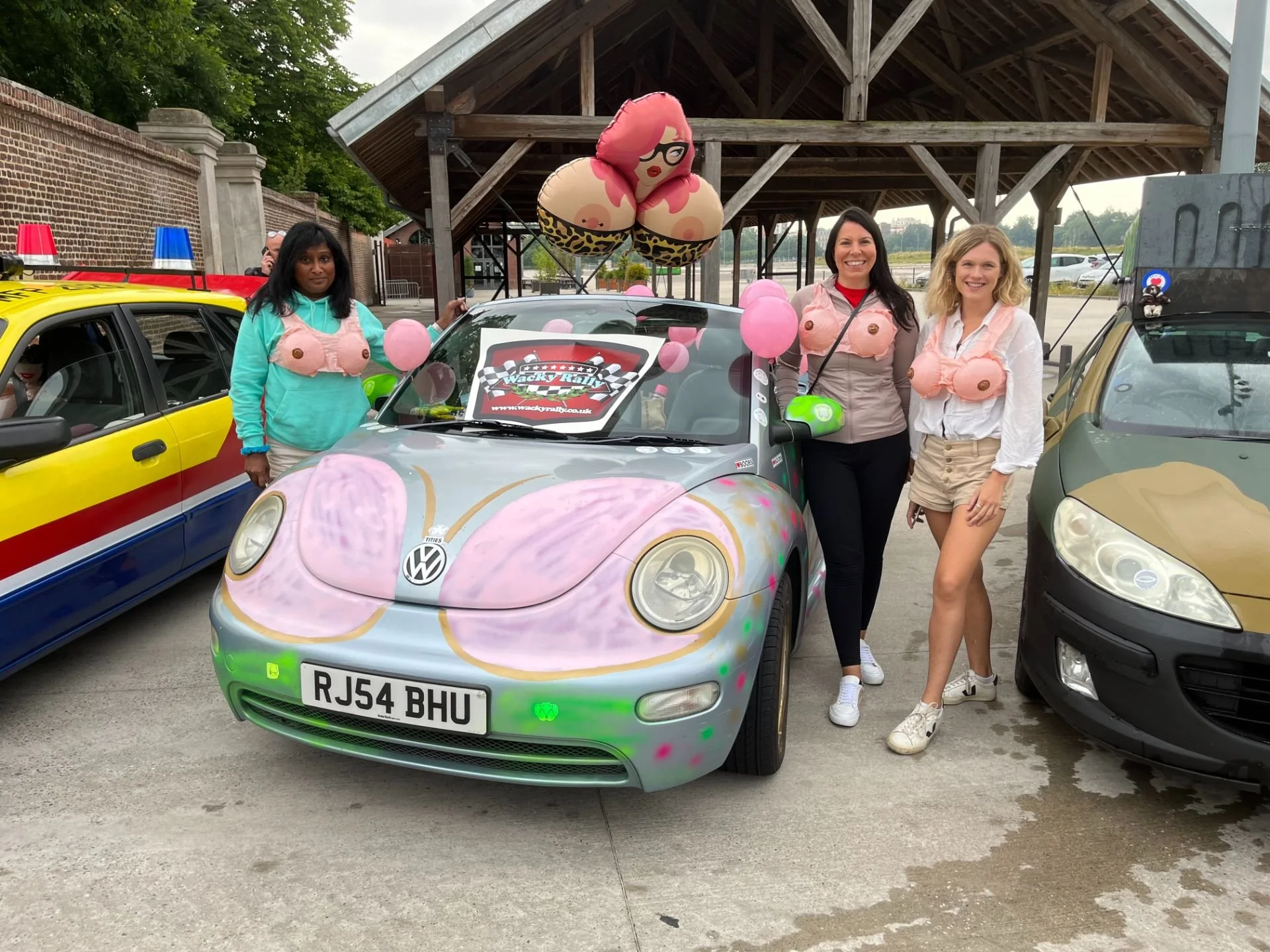 Three women standing next to a decorated Volkswagen car with pink balloons and a large bubble-themed balloon. The women are wearing tops that resemble breasts with nipples. The scene appears to be at a car event or rally, with other painted cars visi