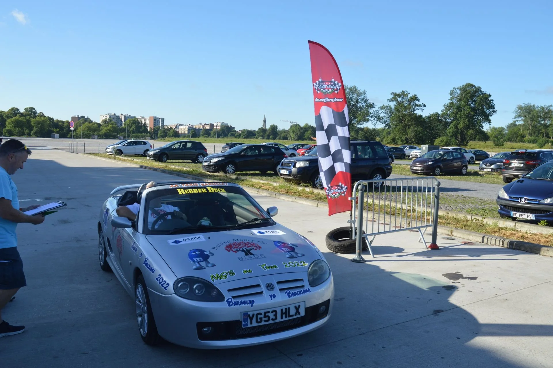 A white convertible car decorated with various colorful stickers and slogans, parked outdoors near a checkered flag banner and metal barrier. A man is standing nearby, taking notes. There are other parked cars and green trees in the background.