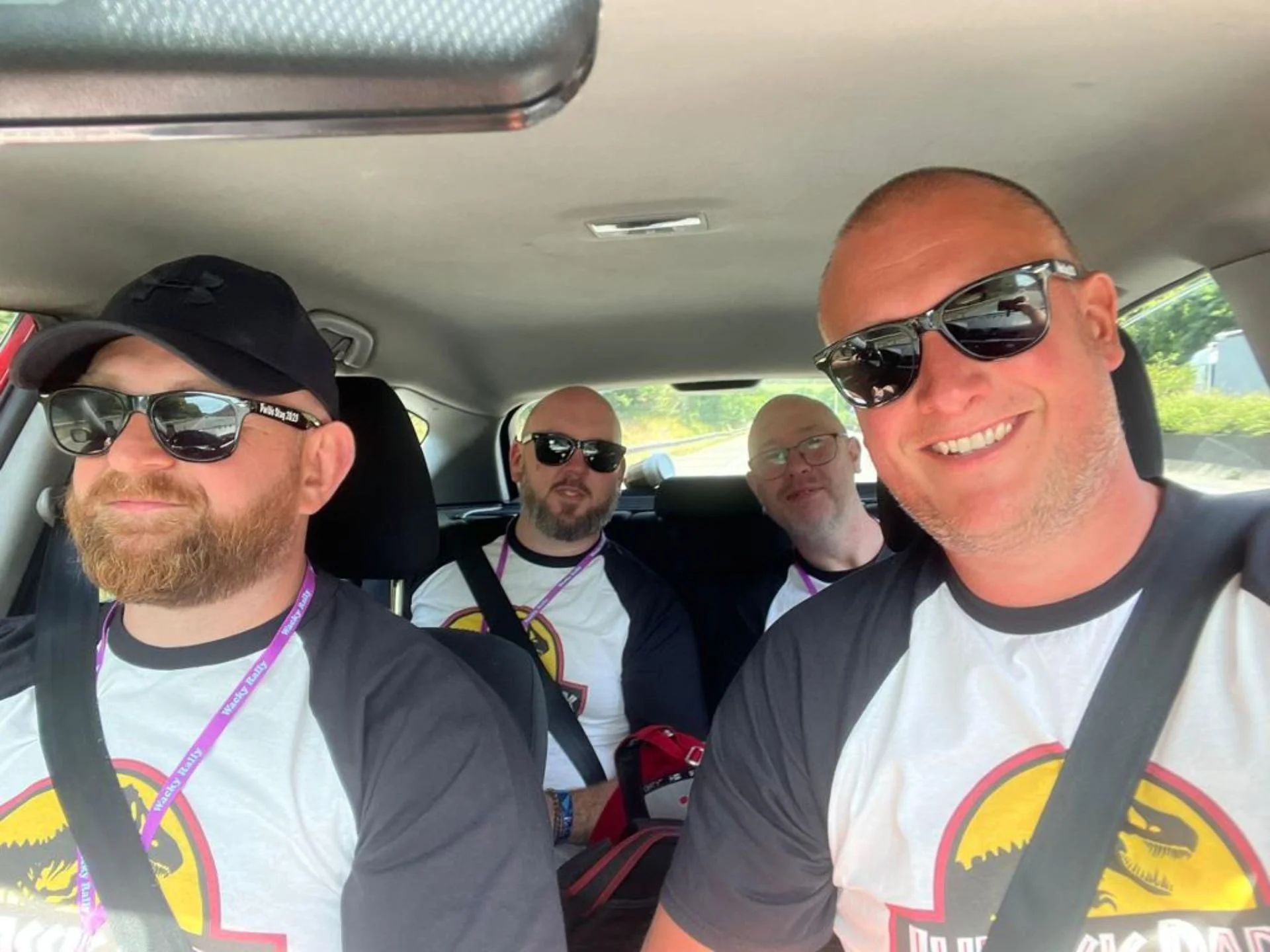 Four men ride together in a car, all wearing sunglasses and matching black-and-white race shirts with a dinosaur logo.