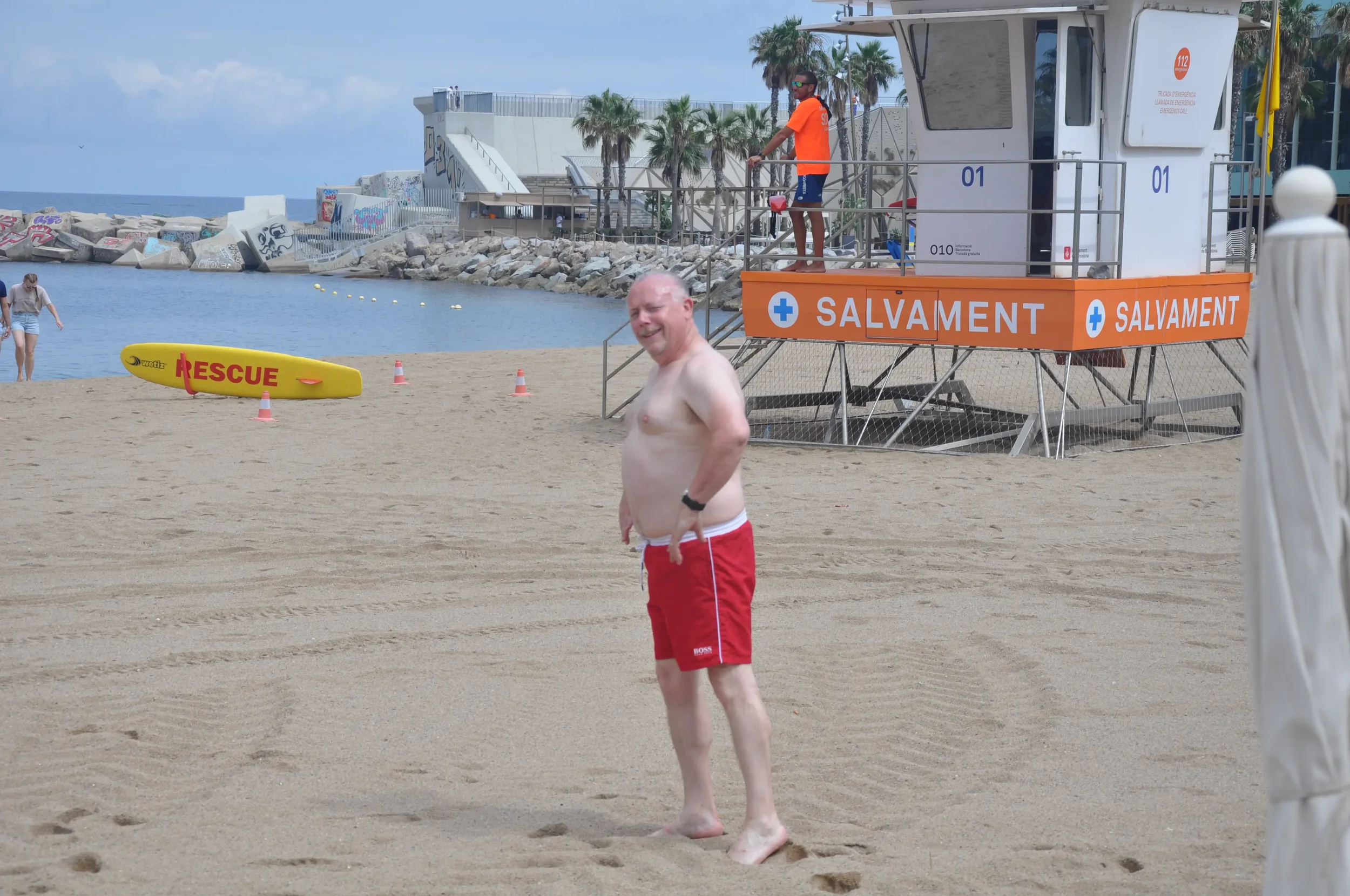 A man in red swim trunks smiling at the beach, with rescue equipment and a lifeguard tower in the background.