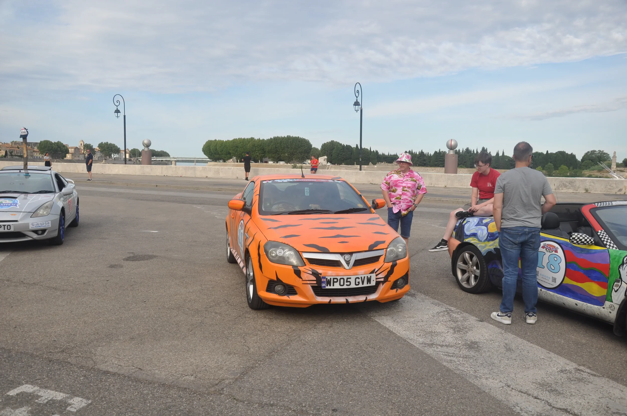 A parking lot with three colorful cars and people standing nearby, with a bridge and trees in the background.