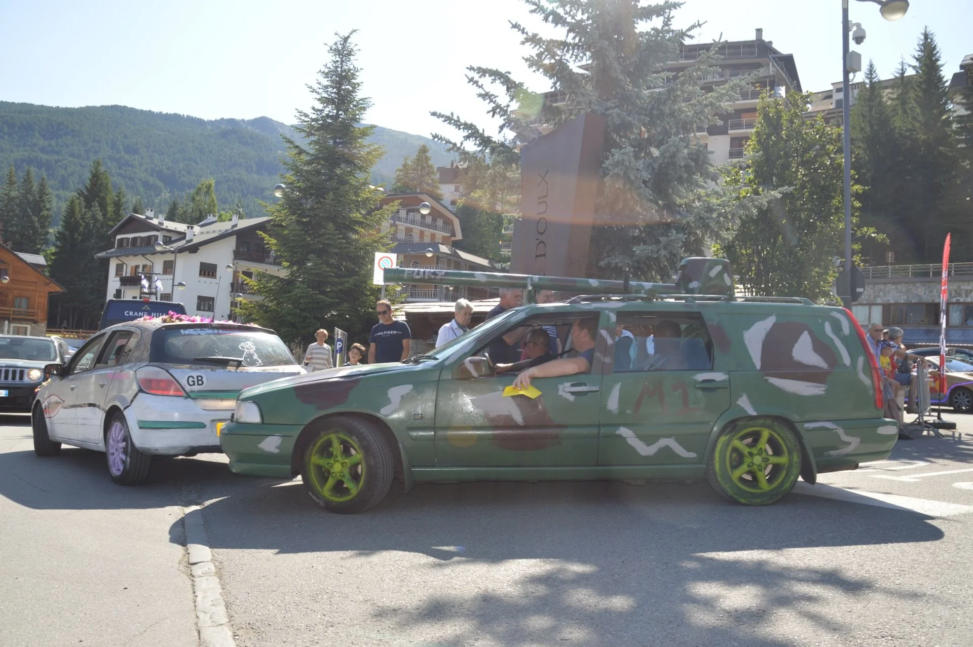 A camouflage-painted station wagon with yellow wheels and a mounted rifle on top, parked in a lot with people walking around, in a mountain town with trees and buildings in the background.