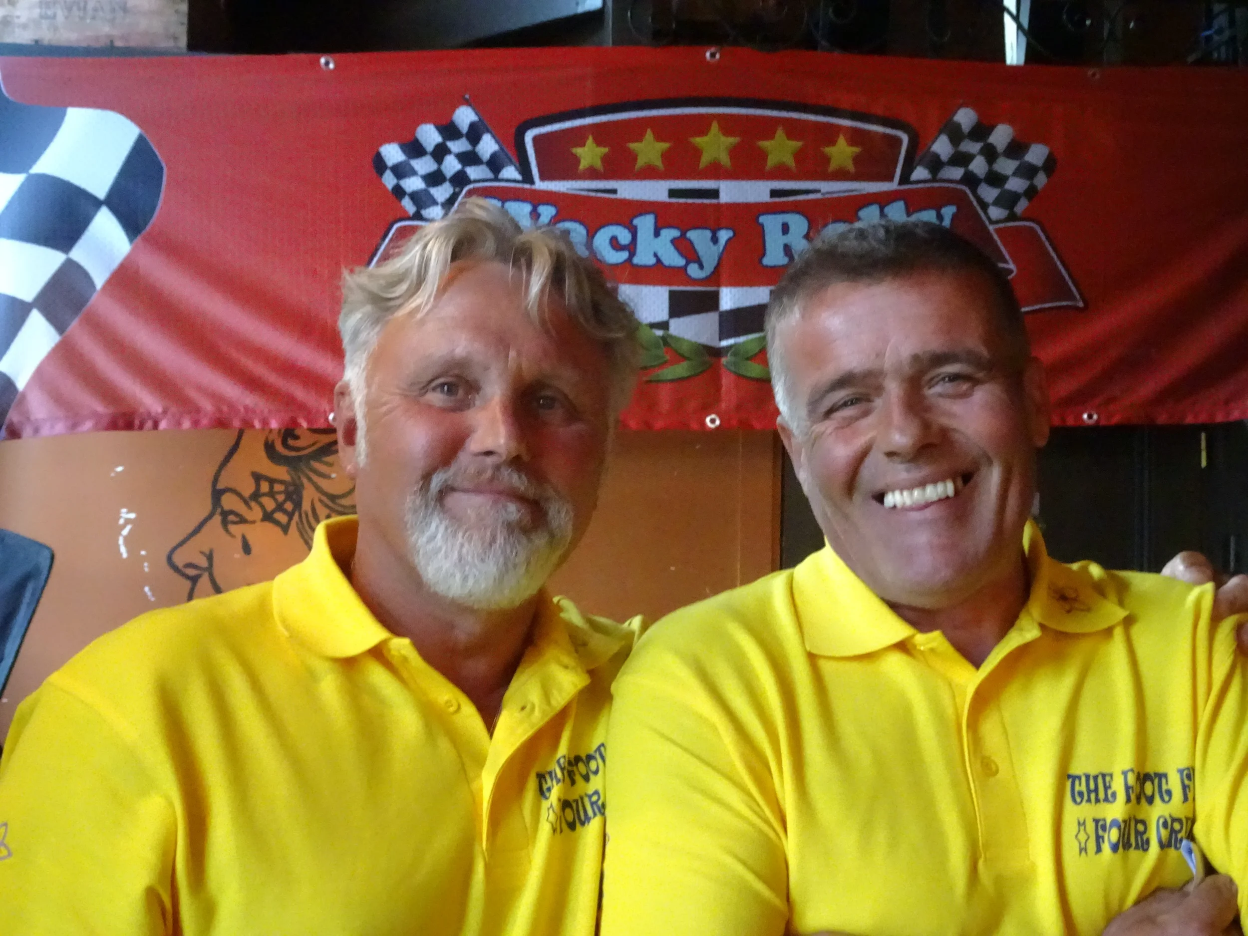 Two smiling men in yellow polo shirts are posing in front of a red banner with racing flags. One man has gray hair and a beard, the other has short brown hair. The banner says 'Rocky R' and has checkered flags on either side.