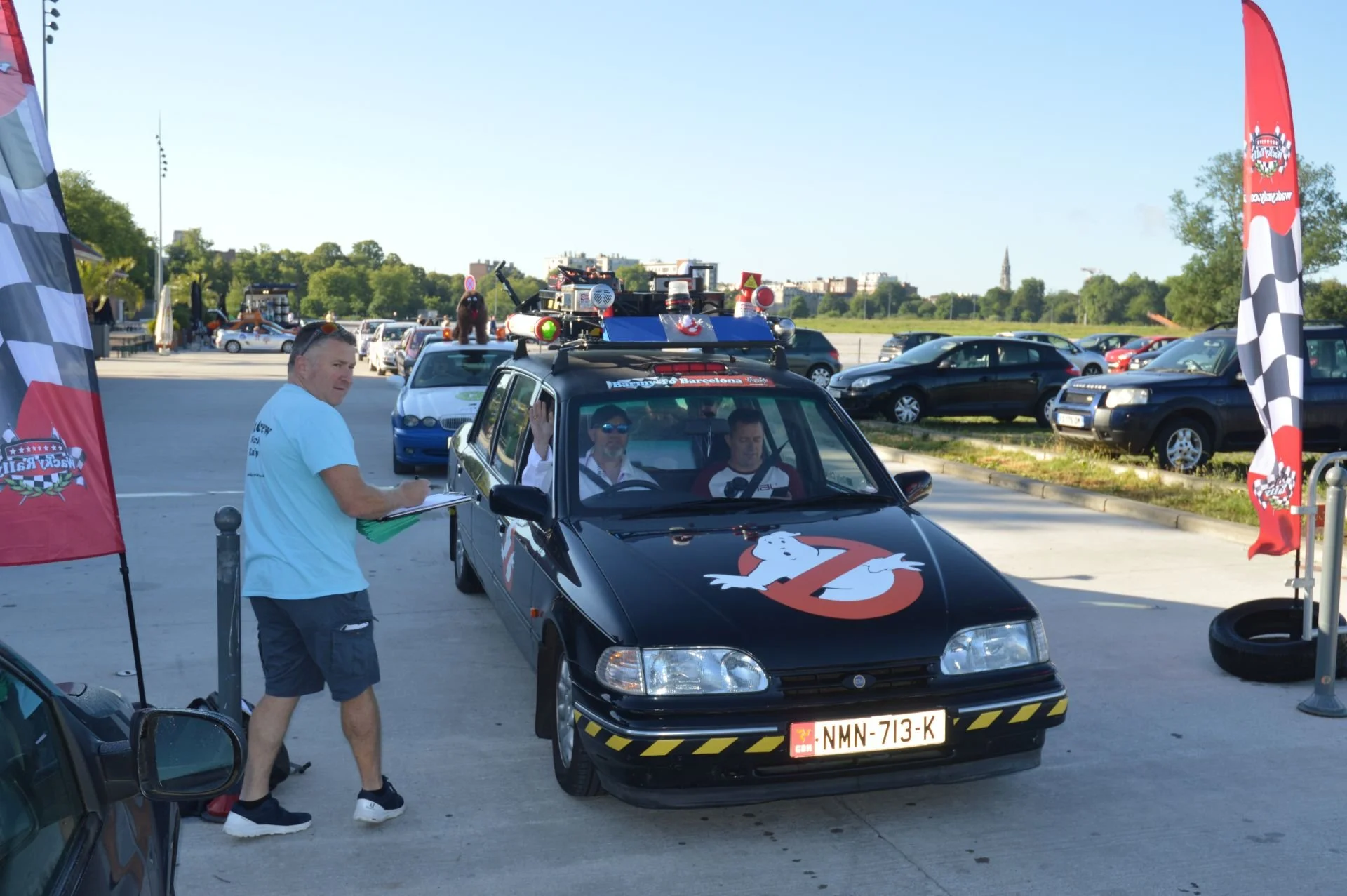A black car with the Ghostbusters logo on the hood, parked in a lot with several other vehicles, at an event with flags. Two men are inside the car, one wearing sunglasses. A man outside the car is holding a clipboard and taking notes.