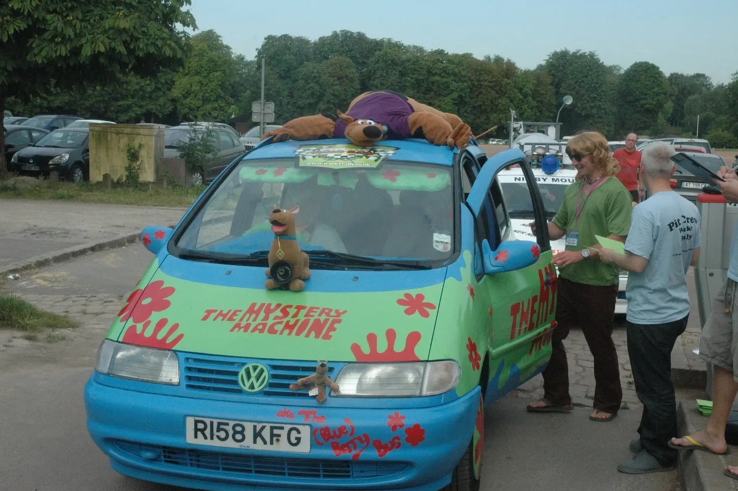 Colorful van decorated with flowers and the words "The Mystery Machine". Cartoon character Scooby-Doo is on the hood, and a man is sitting inside. A woman and a man are talking next to the van, which has someone lying on the roof dressed as Scooby-Do