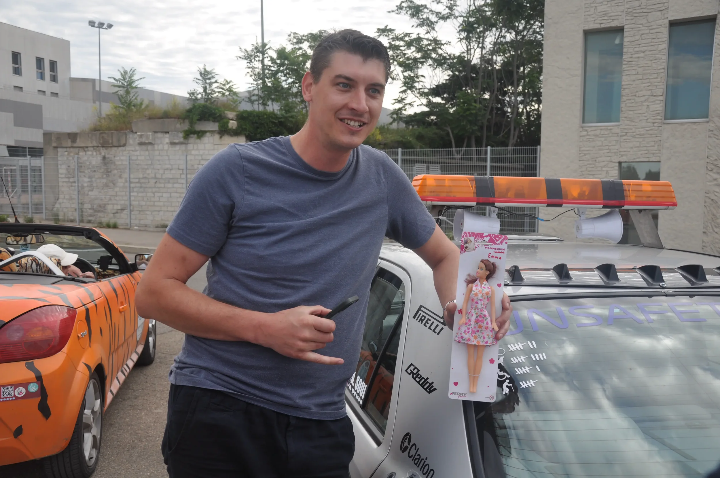 A man is standing next to a police car decorated with a toy doll hanging on the windshield, with another car painted with animal stripes in the background. The man is smiling and holding a phone.