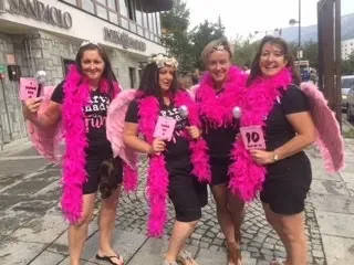 Four women dressed in pink feather boas, angel wings, and black clothing, holding pink signs and participating in a walking event on a city street.