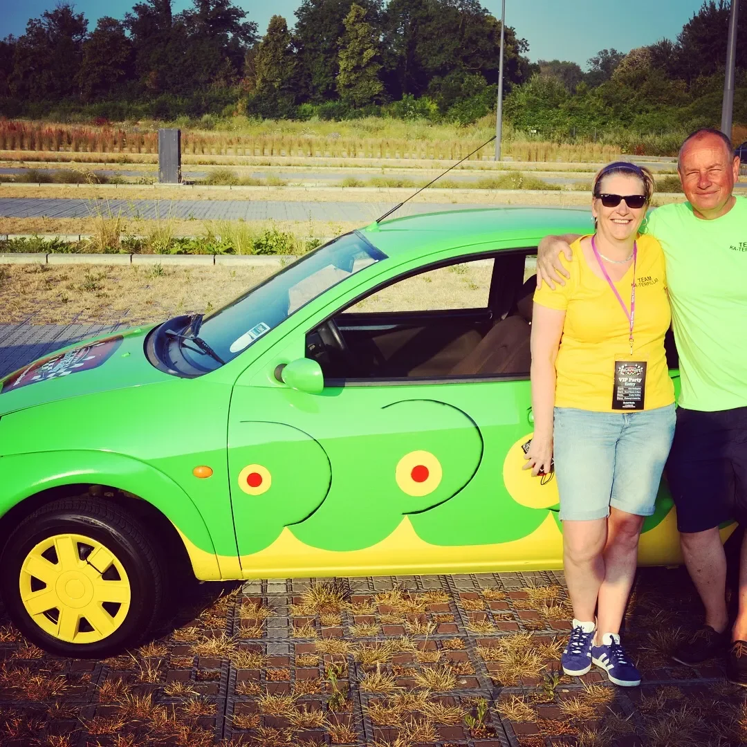 Two people standing next to a bright green car with yellow wheels and a decorative design of green clouds with yellow and red circles. The woman is wearing a yellow shirt, denim shorts, and sunglasses. The man is wearing a light green shirt and has h