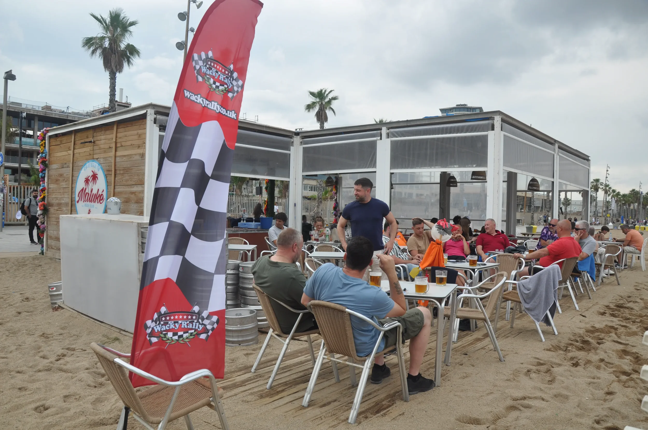 Outdoor dining area on the beach with several people sitting at tables, some with drinks. A man in a navy shirt stands and talks to a group. A red banner with checkered flags and the logo 'Wacky Rally' stands next to a wooden structure with a Malibu 