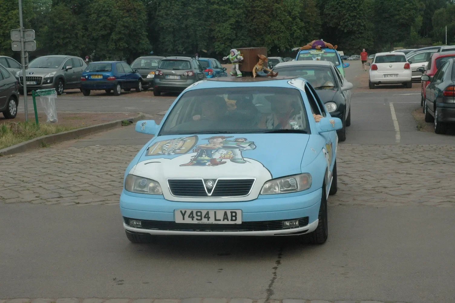 A light blue car with cartoon characters and a flag illustration on the hood, parked in a lot with multiple other cars, some with puppets and toys on top.