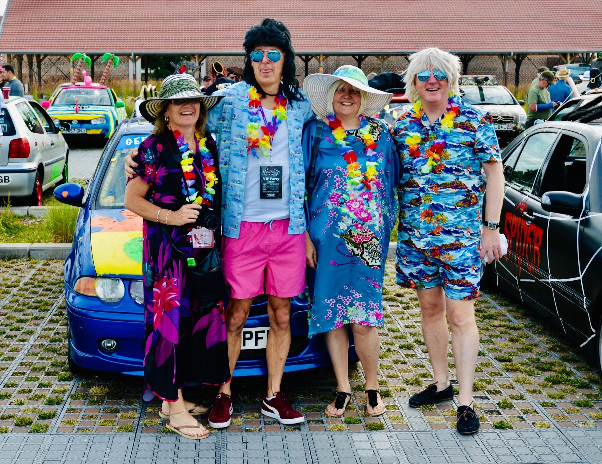 Four people dressed in colorful, tropical-themed clothing and accessories, standing in front of a decorated car at a fun outdoor gathering.
