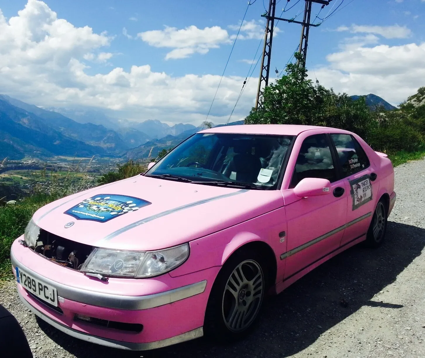 Pink race car parked on a mountain road with a scenic view of mountains and valley in the background.