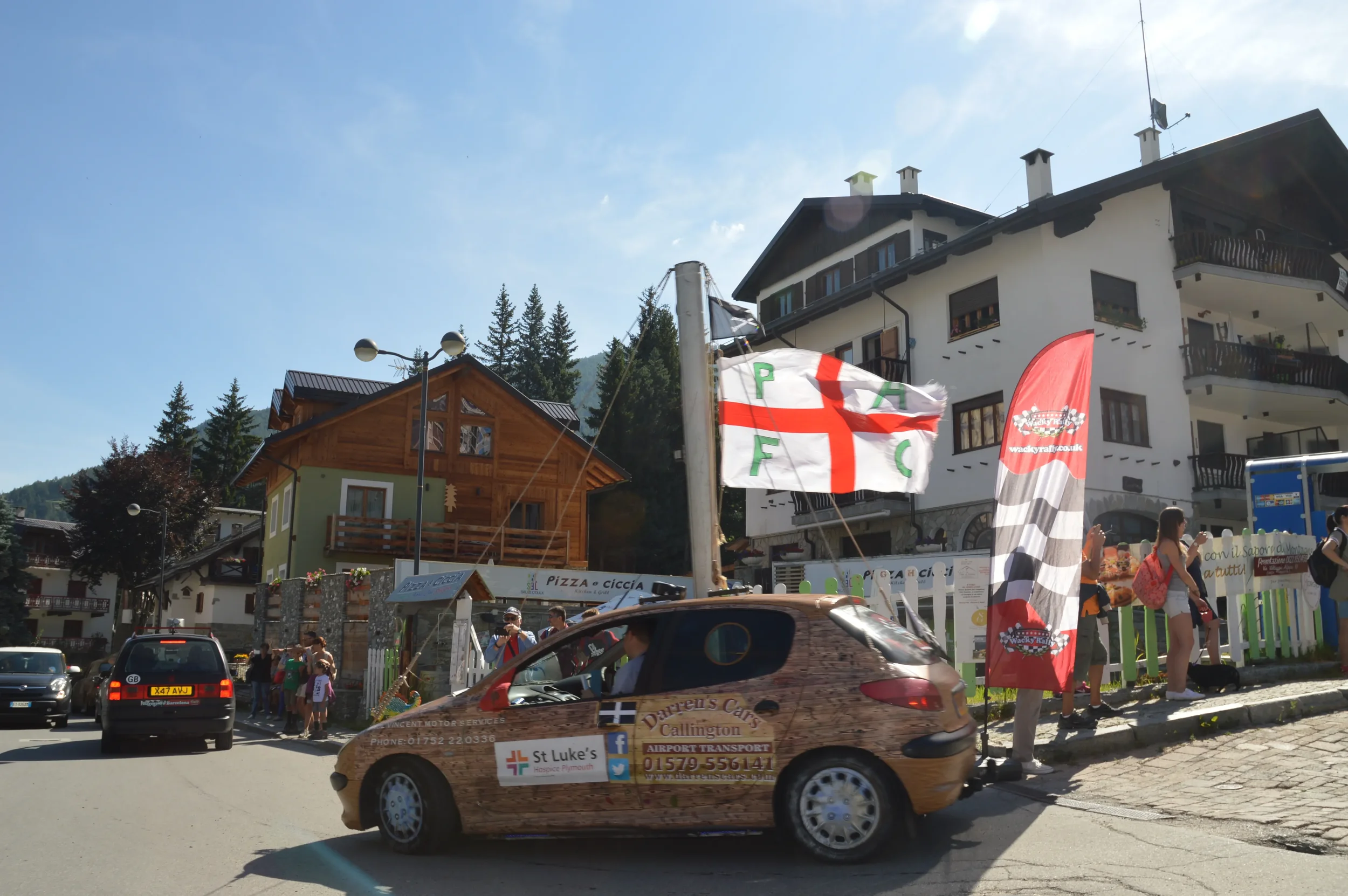 A small town street scene with a decorated car in the foreground featuring advertising for workplaces and a popular logo. People are gathered along the sidewalk, some taking photos. Buildings with balconies and signs are visible in the background und