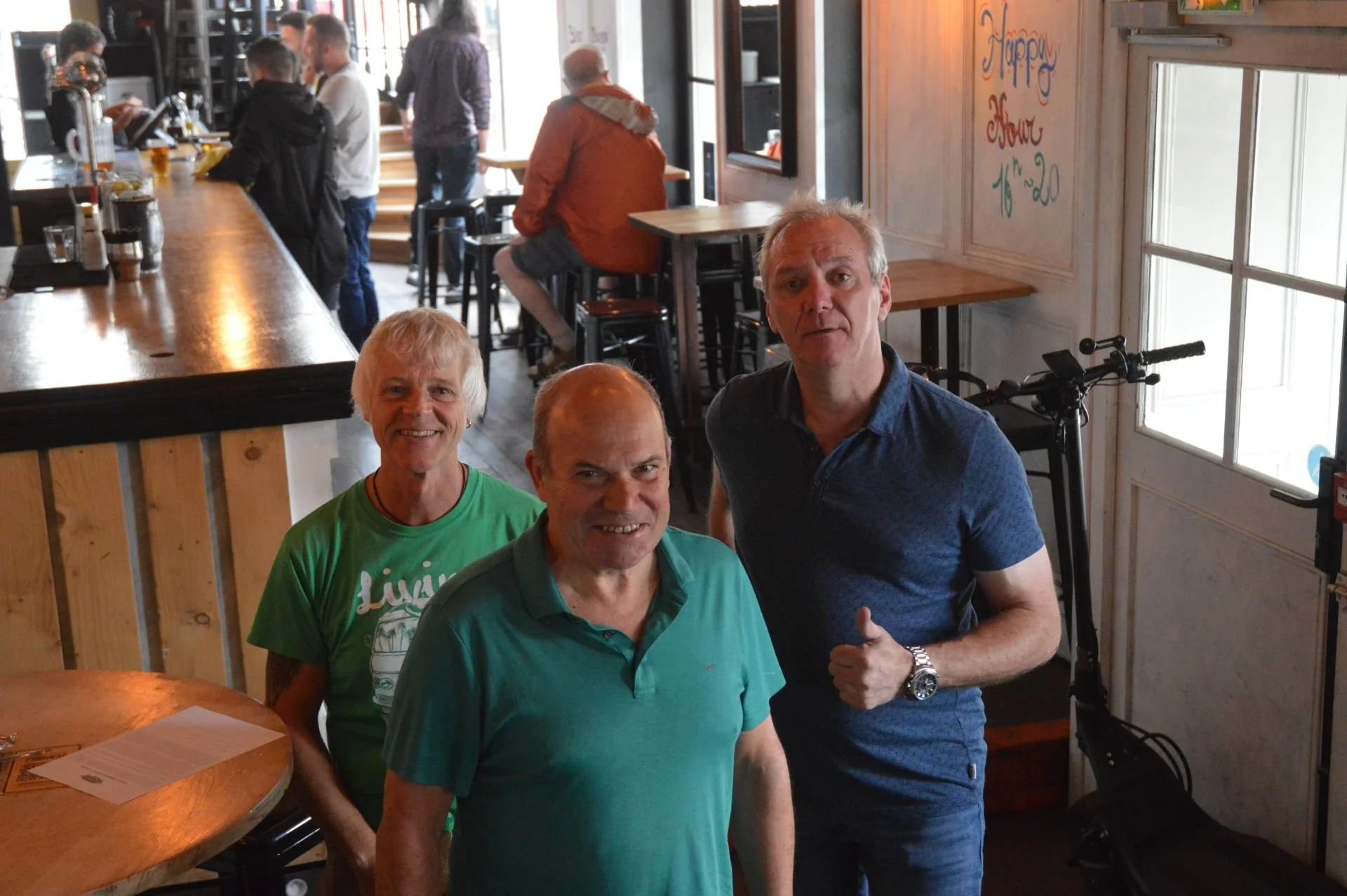 Three men smiling and posing inside a restaurant or cafe, with one man giving a thumbs-up. The background shows other patrons, tables, and a wooden wall with colorful handwriting that says 'Happy Hour 16-20'.