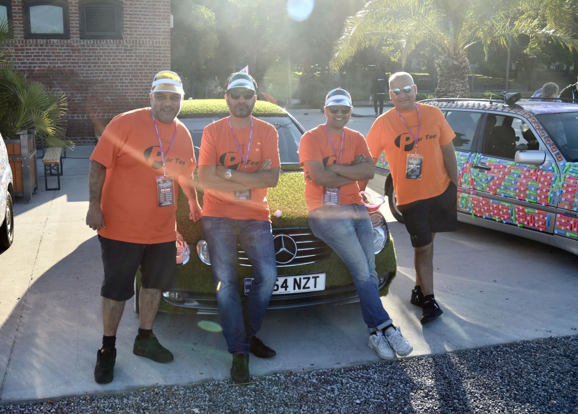 Four men wearing orange T-shirts and sunglasses posing in front of a car decorated with grass and with a license plate reading '154 NZT'. They stand in a parking lot with trees and a brick building in the background, along with a decorated car to the
