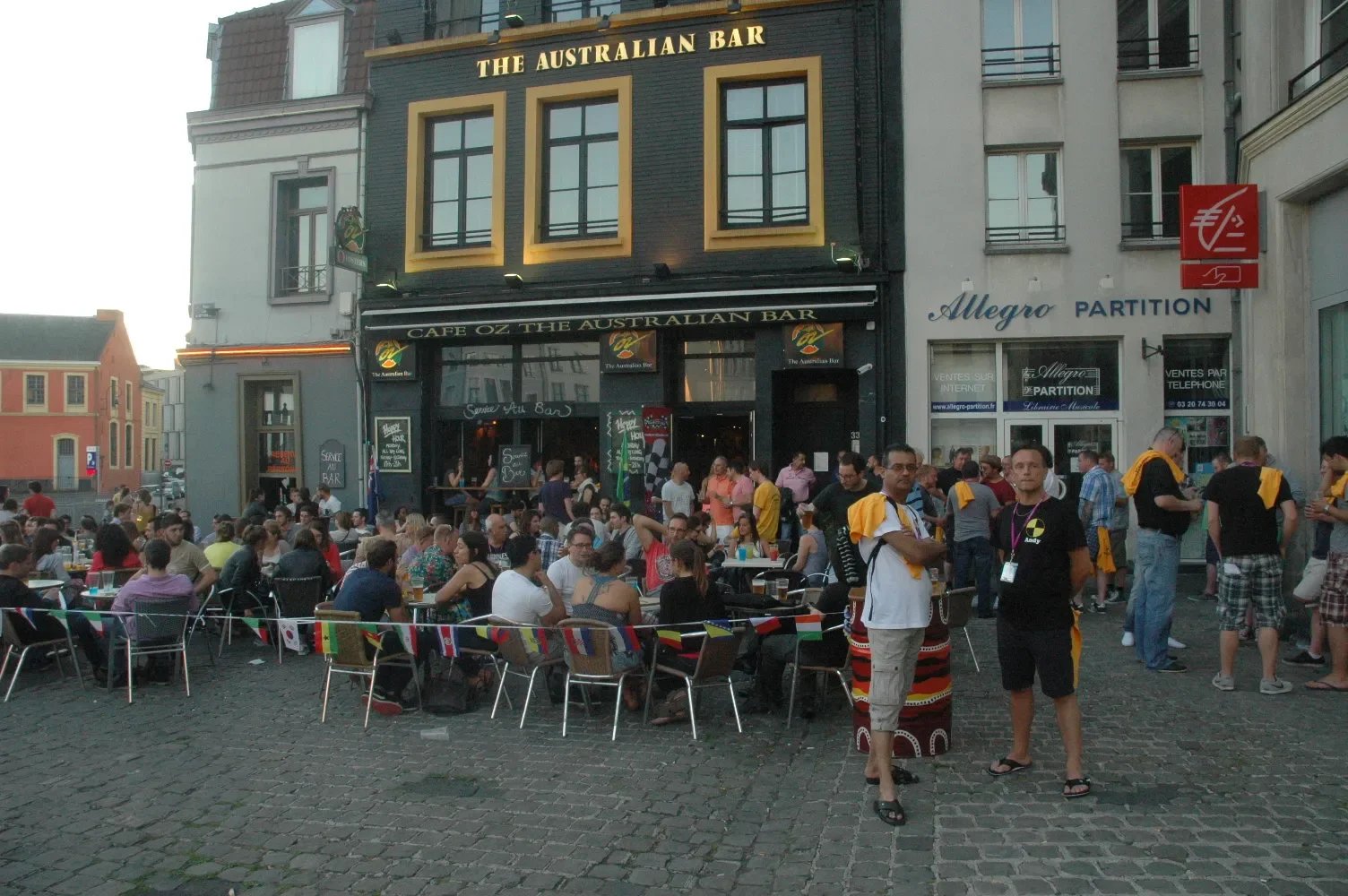 People gathered outside Café Oz The Australian Bar, some sitting at tables and others standing, on a cobblestone street in front of a building with signs and windows, during the evening.