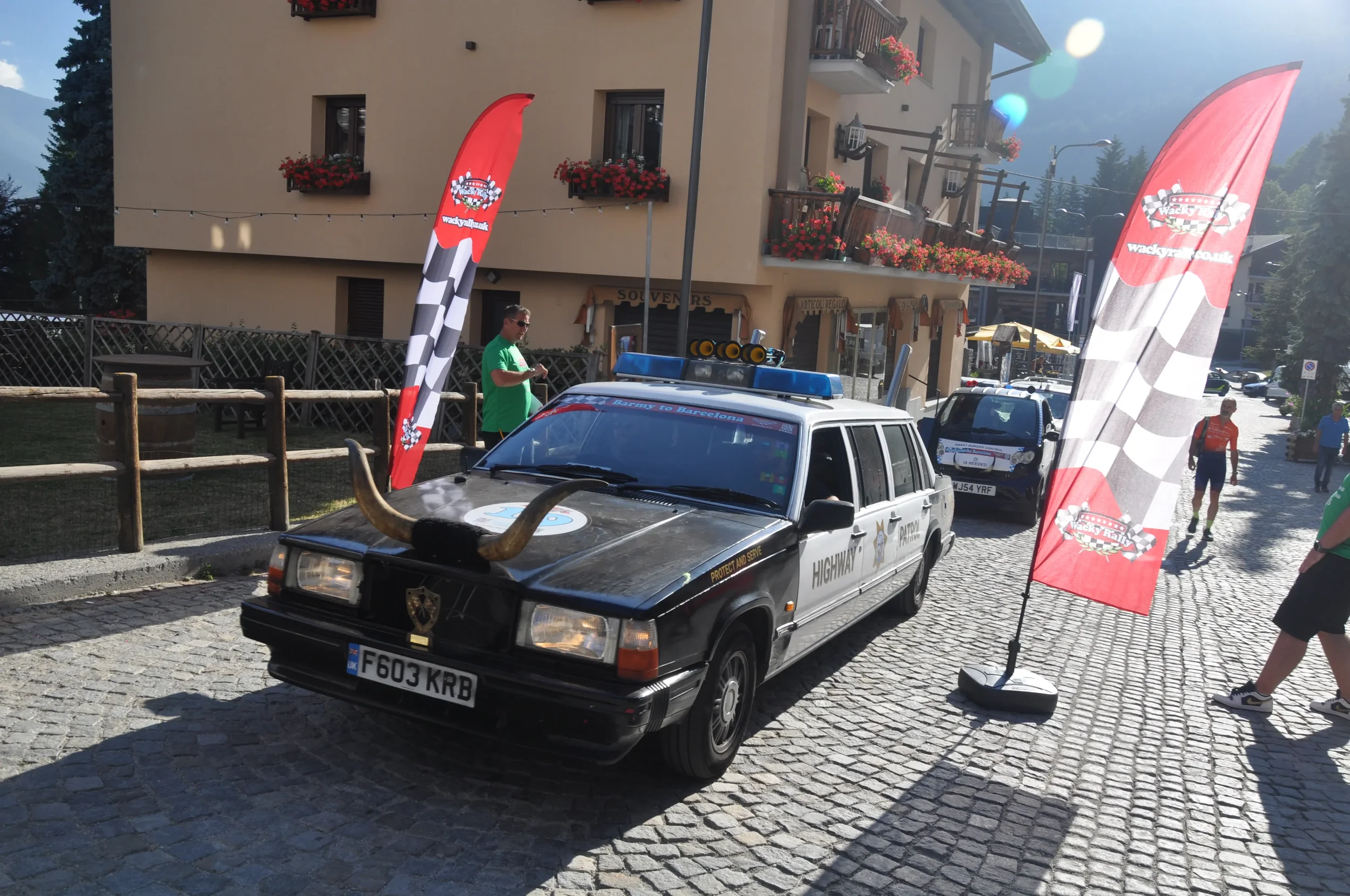 A black and white police car with large horns mounted on the hood, parked on a cobblestone street, with red and white checkered flag banners on either side. There are some people walking and standing around in a small mountain town with buildings, sh