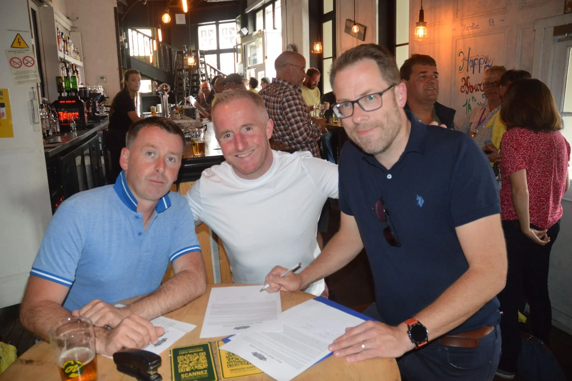 Three men sitting at a table in a bar or restaurant, signing documents with a beer and a mug on the table, and people in the background socializing.
