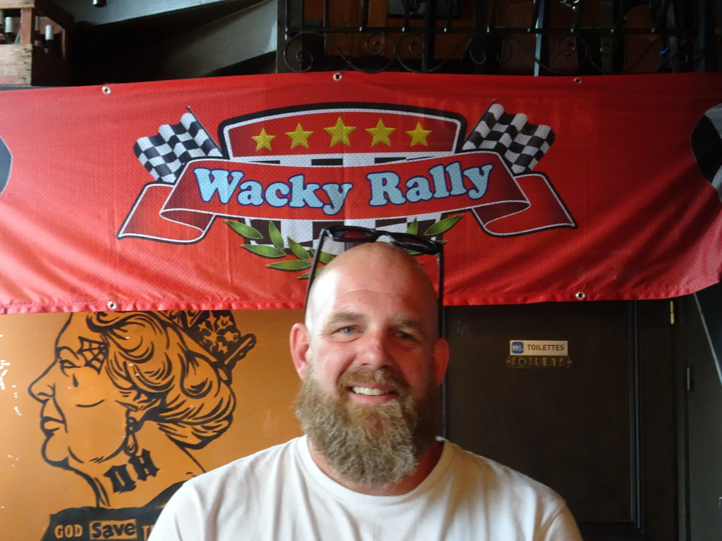 A smiling man with glasses on his head and a beard, standing in front of a red banner that reads "Wacky Rally" with checkered flags, in an indoor setting.