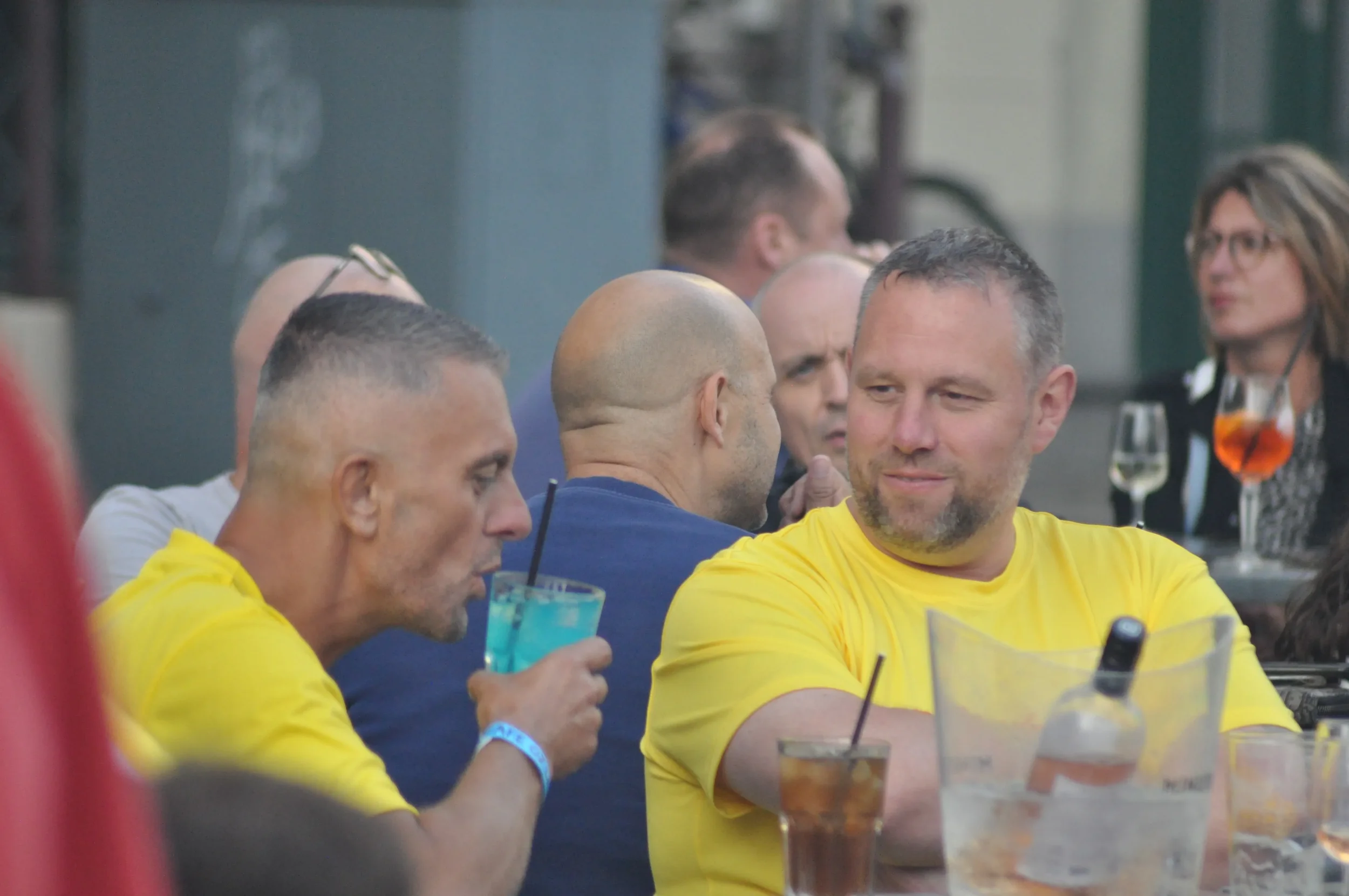 People gathered at a social event, some wearing yellow shirts, with drinks on the table, engaging in conversations.