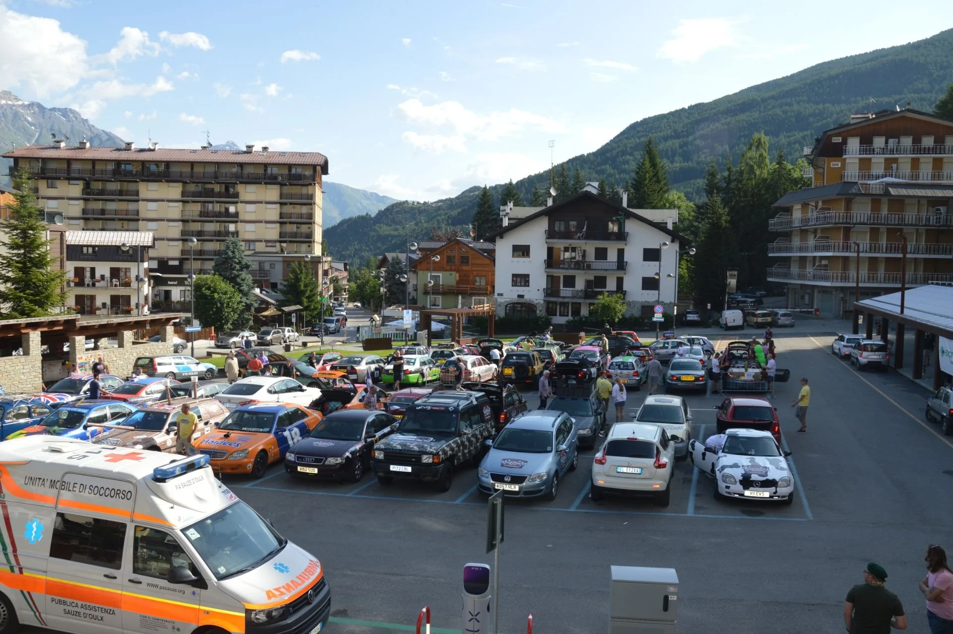 A parking lot filled with various cars, people walking around, and emergency vehicle in foreground, with a backdrop of residential buildings and mountain scenery.