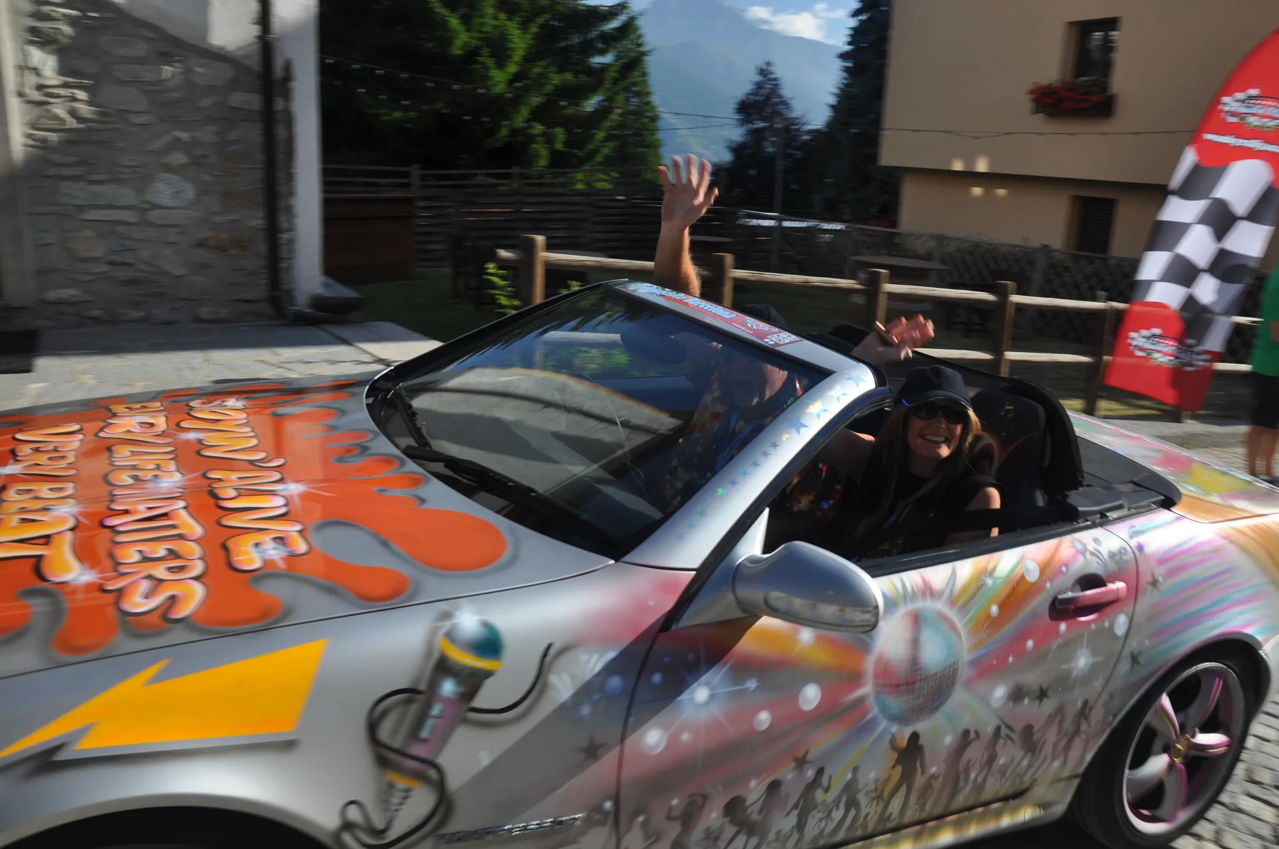 People in a colorful convertible car with artwork and text on the hood, waving and smiling during a sunny outdoor event.