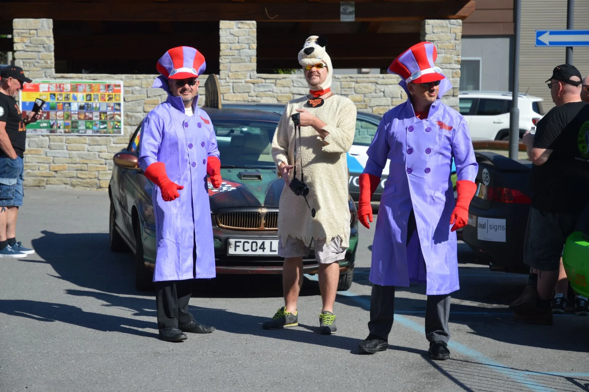 Three people dressed in costumes standing in a parking lot, with a stone building in the background. Two of them are dressed as characters from the TV show 'The Wacky Races,' wearing purple coats and red, white, and blue top hats. The third person is