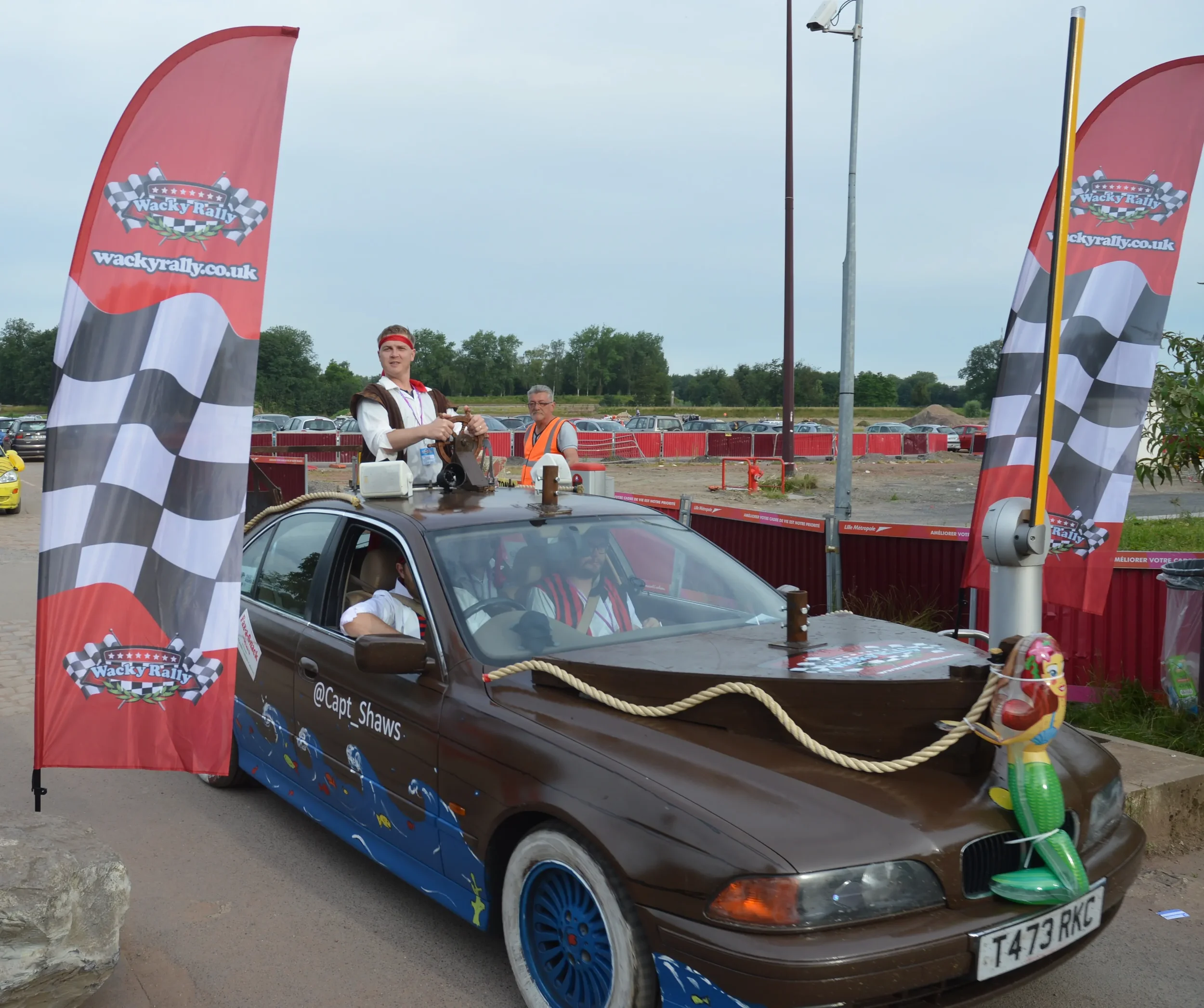 A brown car decorated with nautical-themed paintings and a large inflatable toy on the front, participating in a rally with two people inside and a man in a brown vest and woman in a grey vest on the roof, surrounded by red flags with checkered racin
