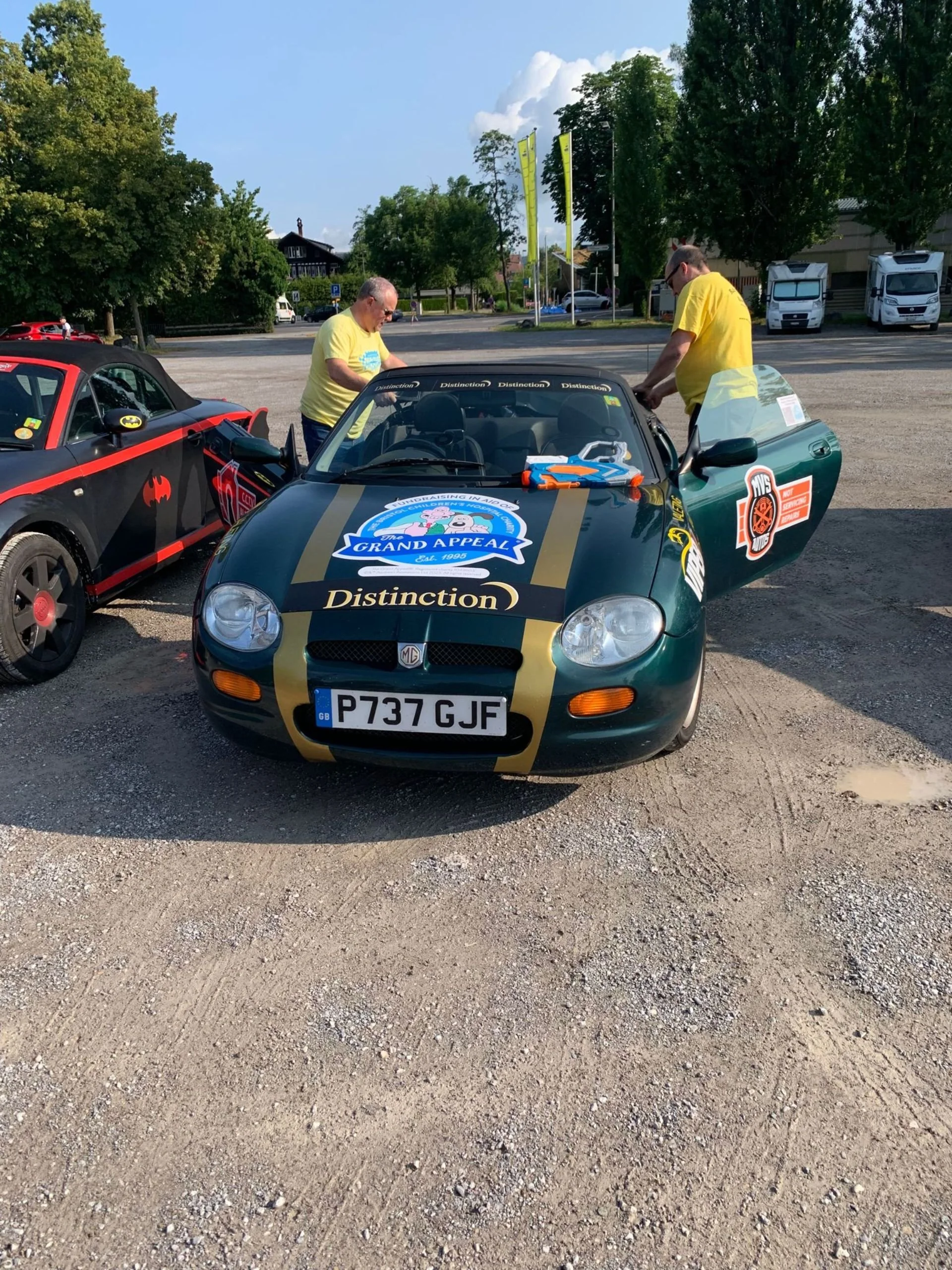A dark green MG sports car with a racing stripe, several stickers, and the word "Distinction" on the hood, parked in a lot with two men in yellow shirts working on it. Another car with Batman and other stickers is parked beside it.