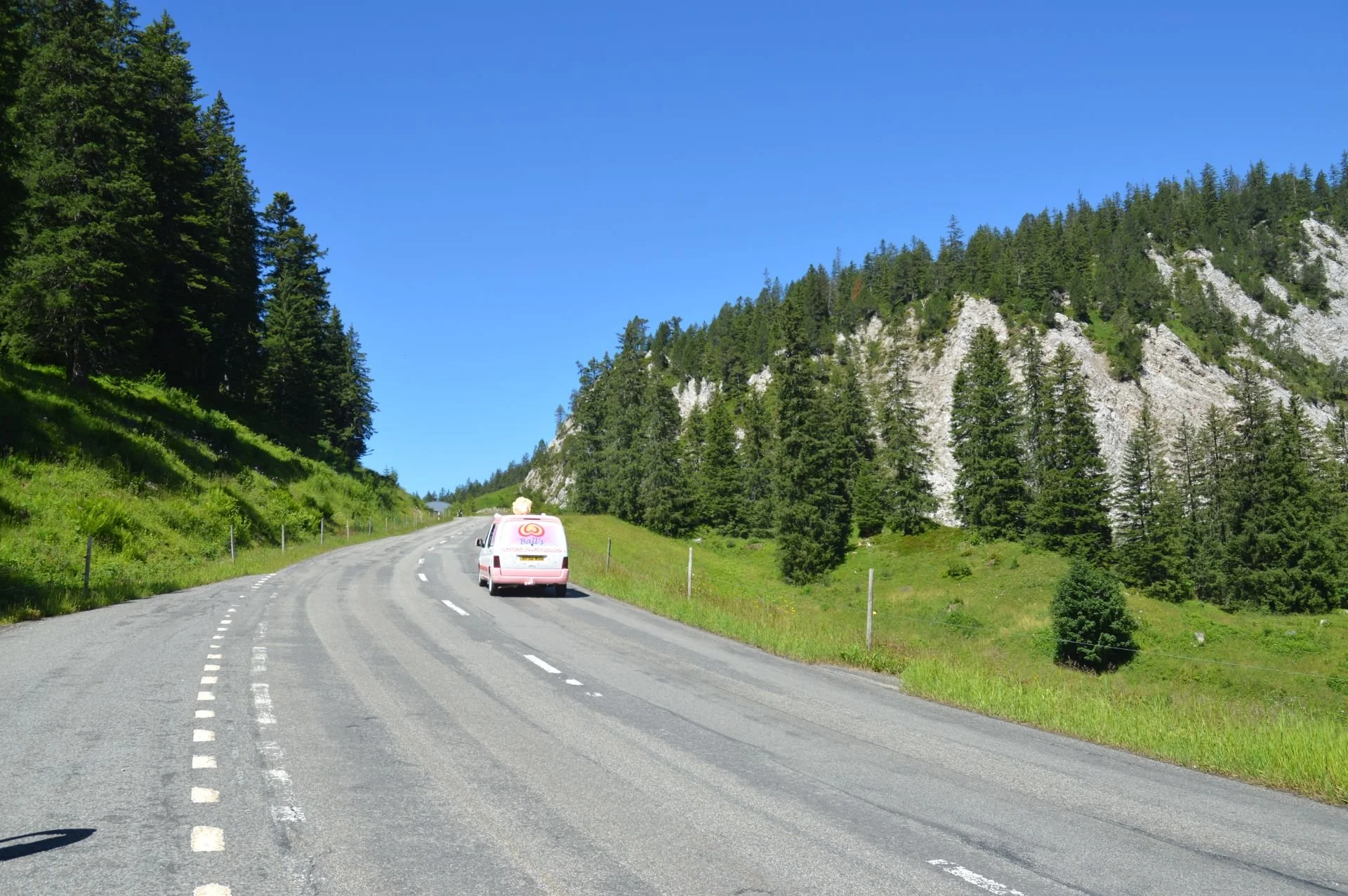 Pink ice cream truck driving on a winding mountain road surrounded by green trees and rocky hills under a clear blue sky.