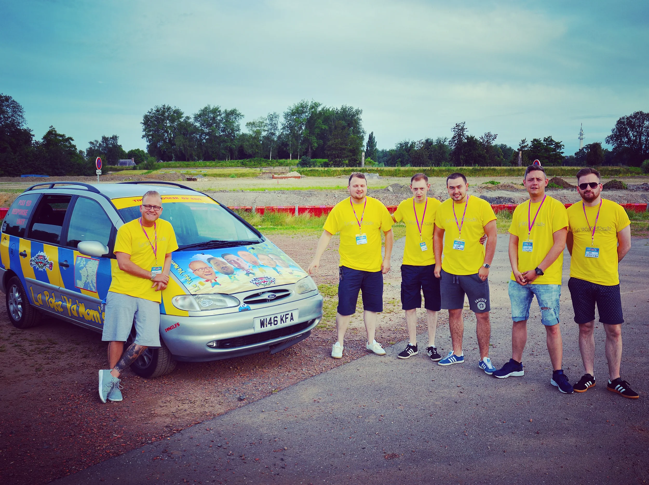 Group of six men standing next to a car with cartoon graphics, all wearing yellow shirts, outdoors on a cloudy day.