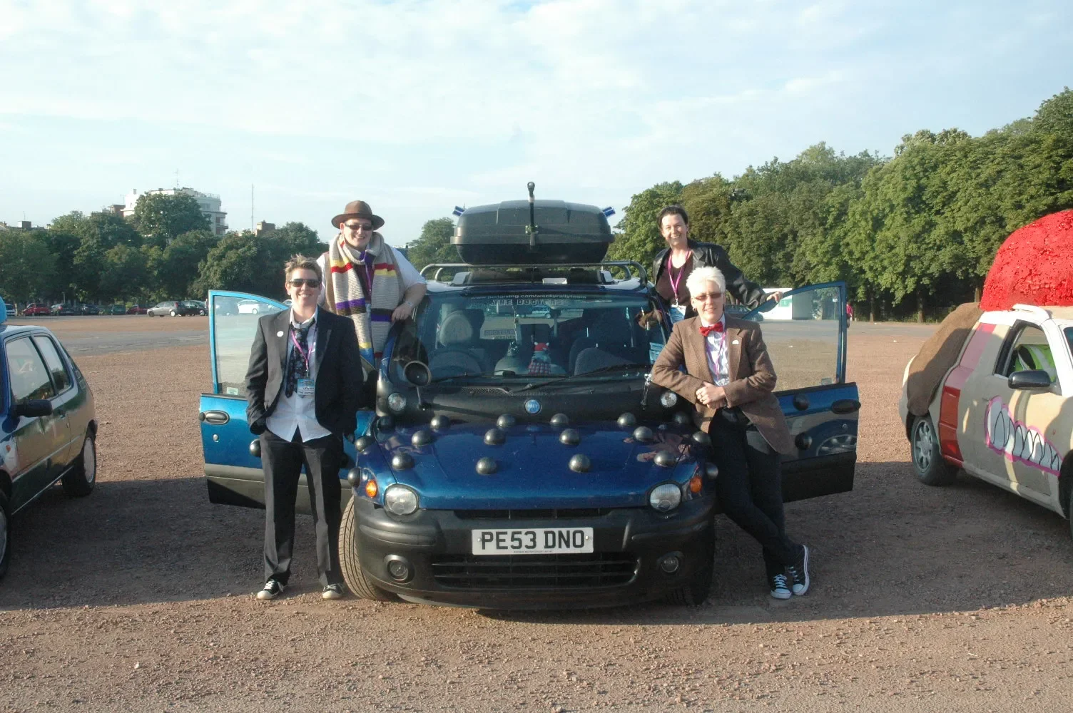 Group of five people posing around a decorated black car with the license plate 'PE53 DNO' in an outdoor parking lot, with two other decorated cars nearby, trees, and a building in the background.