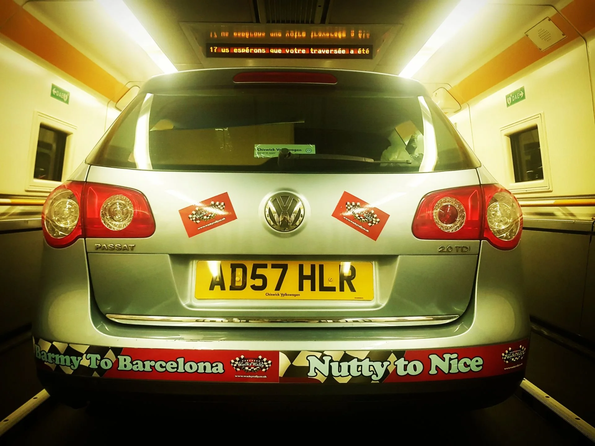 Rear view of a silver Volkswagen Passat with a yellow license plate, admission stickers on the back, and red bumper stickers that say 'Barmy To Barcelona Nutty To Nice' with checkered flags.
