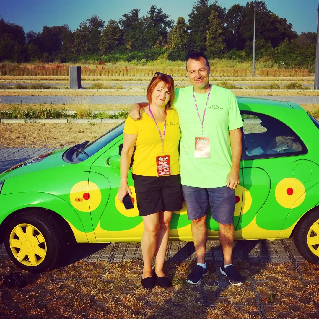 A man and a woman standing next to a brightly decorated green car outdoors during daytime. They are smiling and wearing event lanyards, with a background of trees and open space.