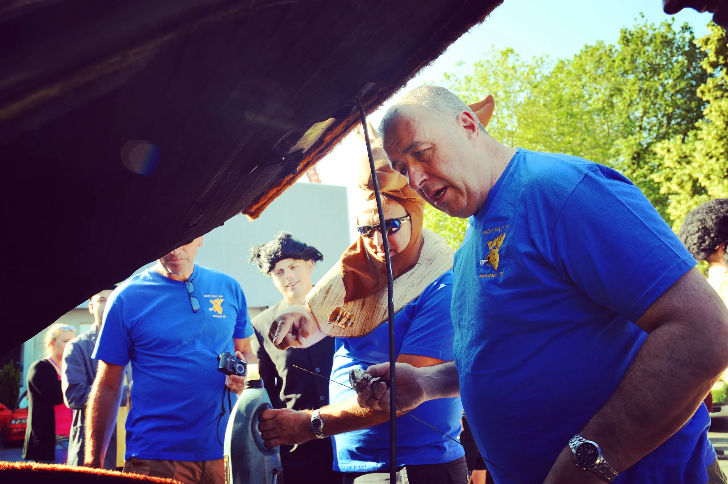 A group of people, some in costumes, gathered outdoors under a large object. One man in a blue T-shirt appears to be working on something with a tool, while others observe. The setting is sunny with trees in the background.