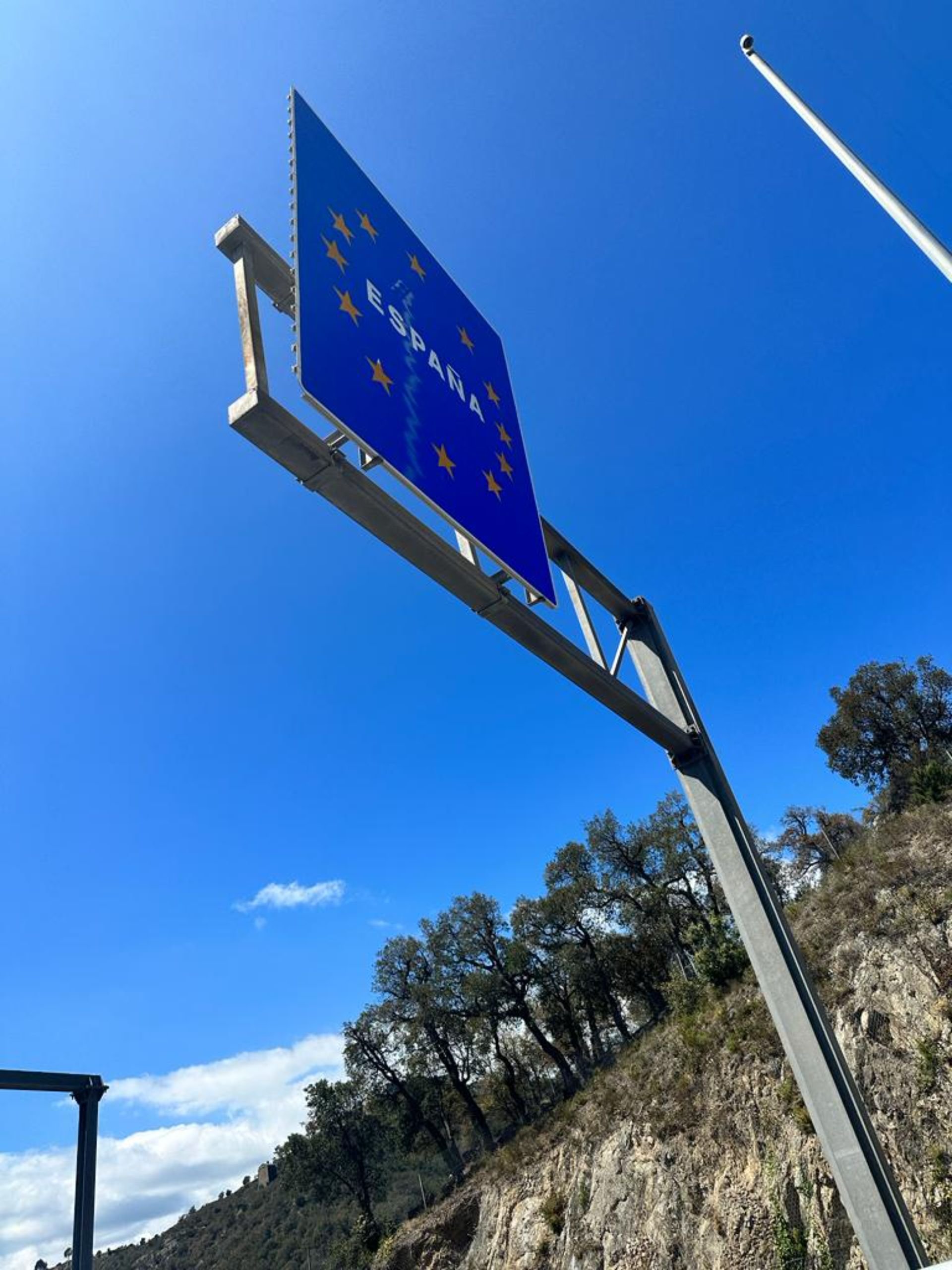 Blue road sign with yellow stars and the word 'ESPAÑA' indicating entry to Spain, against a bright blue sky and hillside with trees.