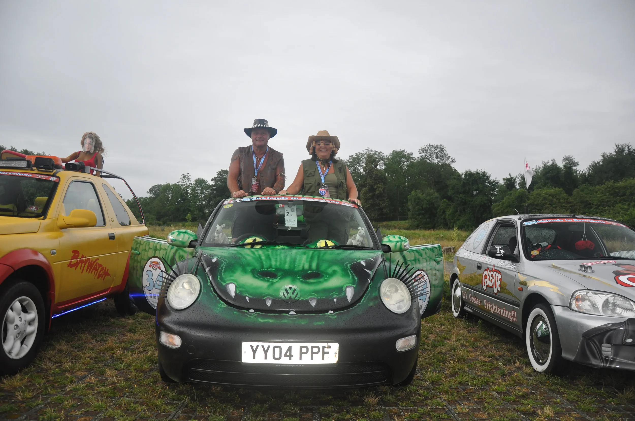 Two people standing through the sunroof of a black Volkswagen Beetle with a green monster face painted on the hood, parked between a yellow car and a silver car with racing decals, outdoors on grassy terrain.