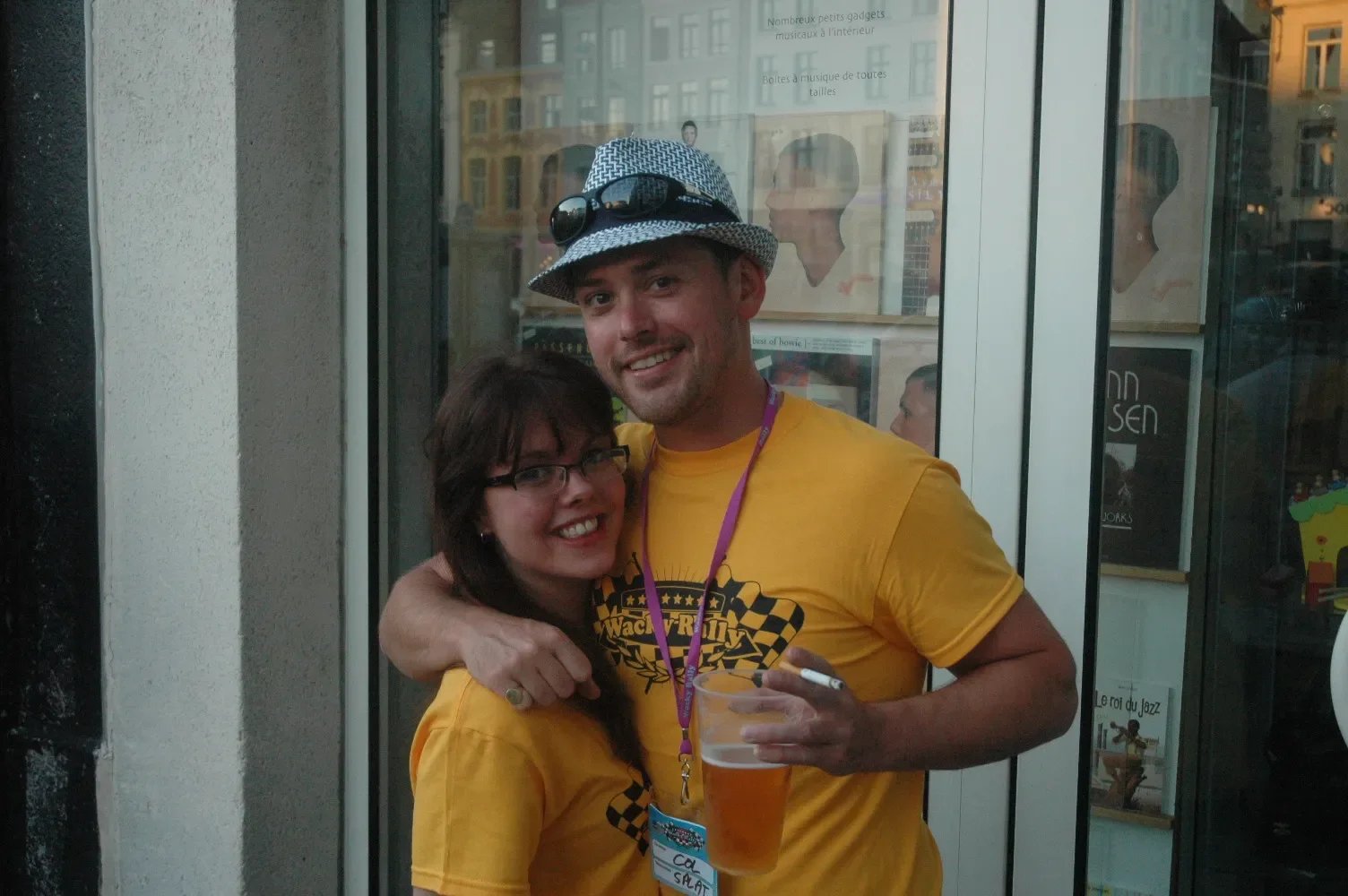 A man and woman standing outside, smiling and hugging, with the man holding a drink in his hand. The man is wearing a yellow T-shirt and a hat with sunglasses, and the woman is wearing glasses and a yellow T-shirt. They are in front of a glass door w