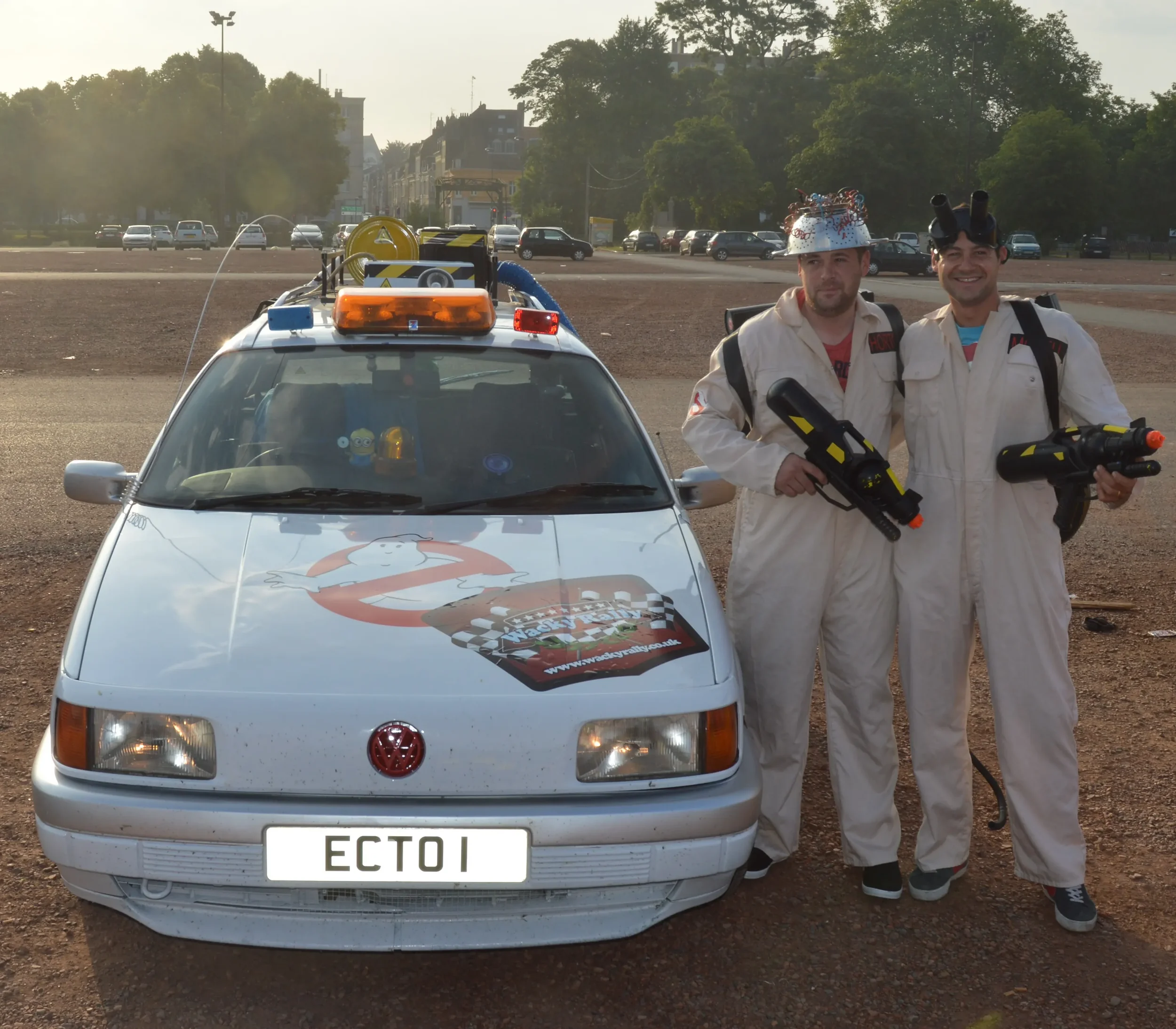 Two people dressed as Ghostbusters characters standing next to a VW Ecto-1 car on a dirt lot during sunset, both holding laser gun props and smiling.