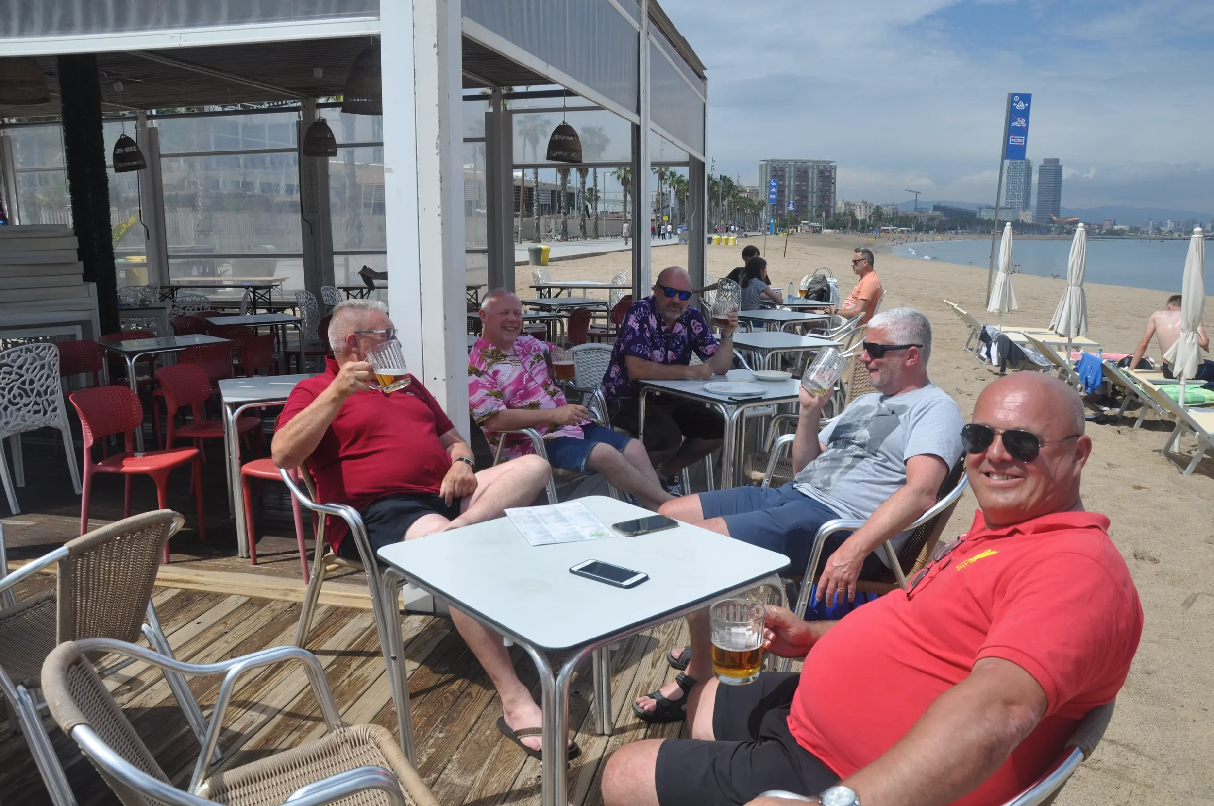 Group of five men sitting outside at a beachside bar, holding drinks and smiling, with the sandy beach, beach umbrellas, and buildings in the background.
