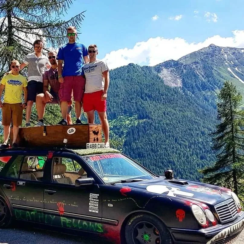 Group of seven people standing on top of a black car with a mountain and forest background during the day.