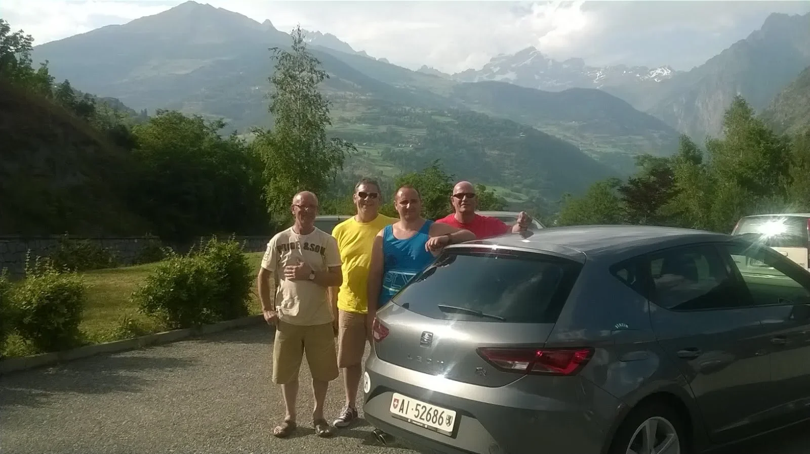Four men standing next to a silver car with a mountainous landscape in the background.