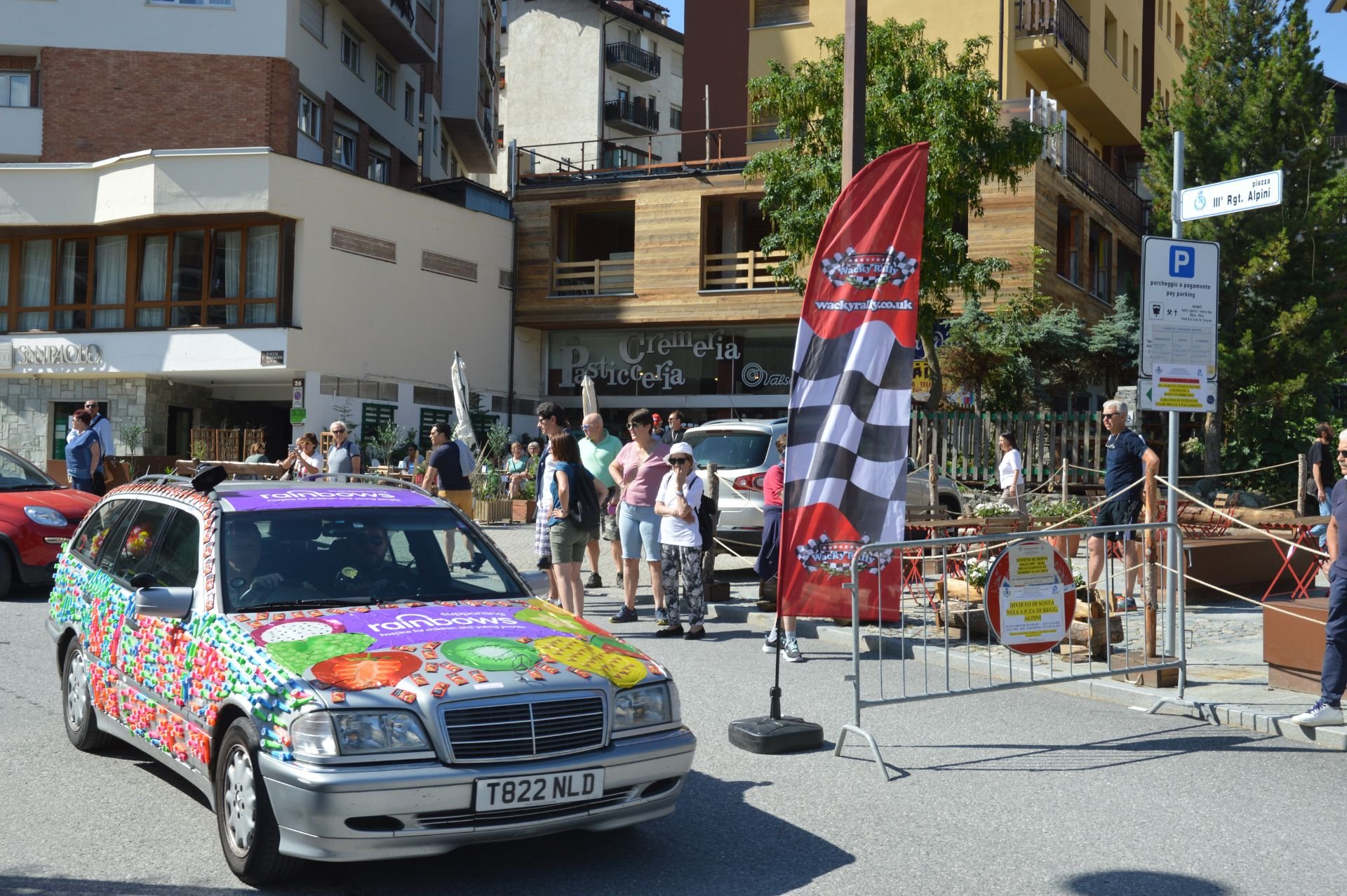 A street scene with a decorated Mercedes-Benz car covered in colorful candy-themed decorations at an outdoor event. People are standing and walking near a barrier with flags, and a sign indicates parking information. Buildings and trees are visible i
