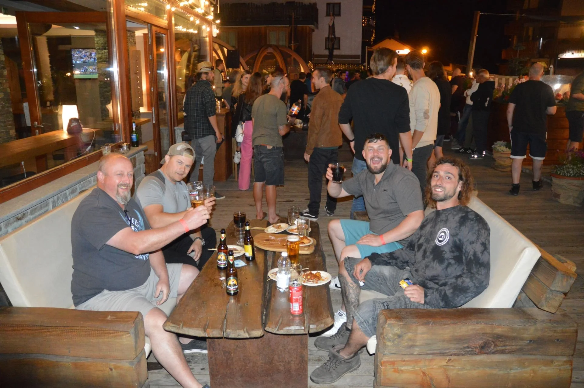 Four men sitting at a wooden table outdoors, enjoying drinks and pizza, with a lively crowd in the background at night.