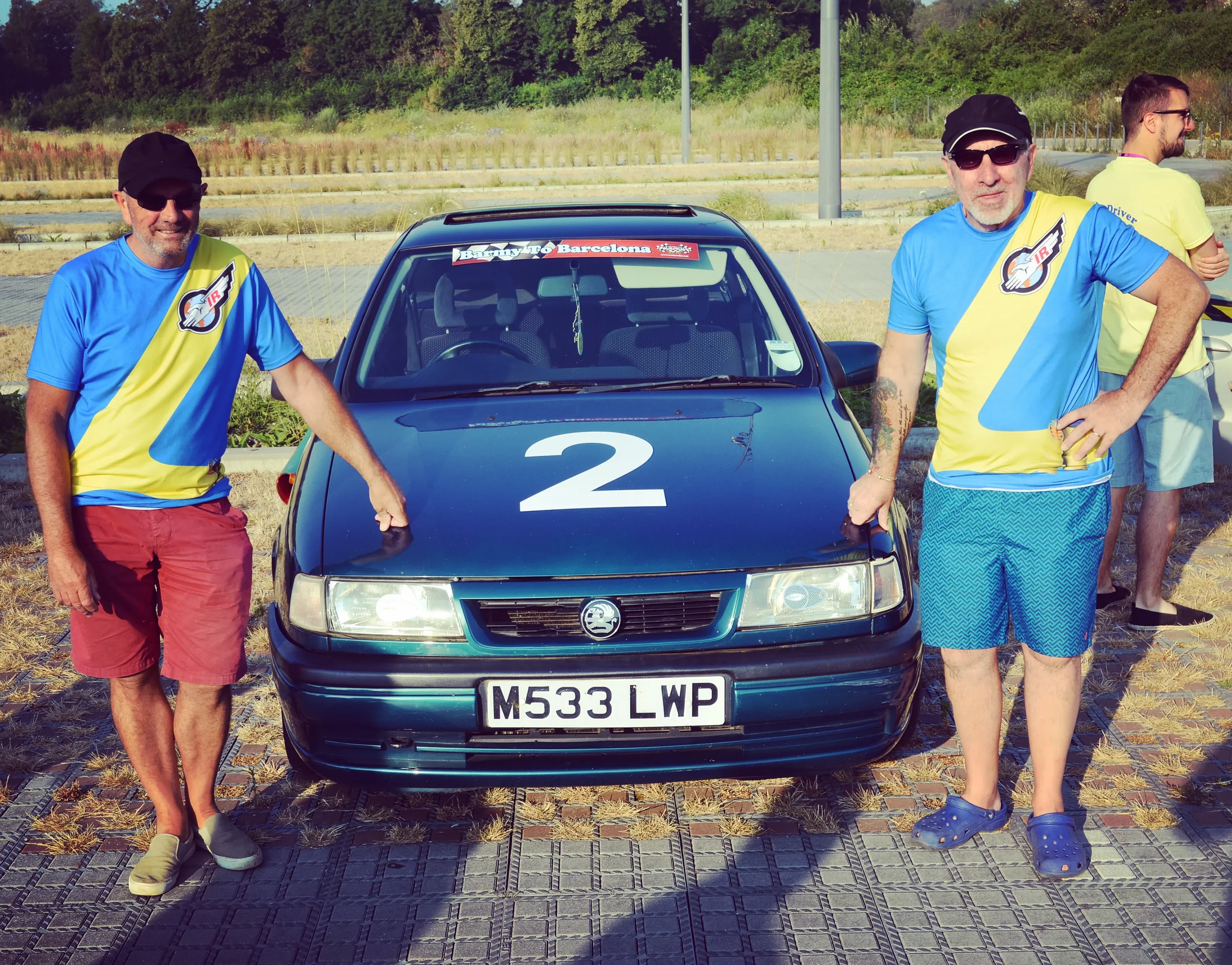 Two men in colorful t-shirts and shorts stand on either side of a black rally car with a large number 2 on the hood. The man on the left is wearing a black cap and sunglasses, and is touching the car. The man on the right is in a black cap, sunglasse