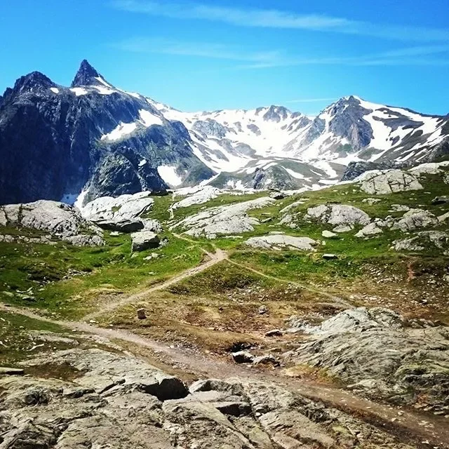 A mountain landscape with rocky terrain, green patches, snow-capped peaks, and a clear blue sky.