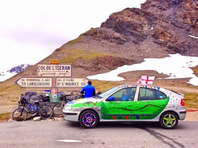 A decorated car with French flags parked on a mountain road, with bicycles and a person in blue nearby, in a rocky mountain area with snow patches.