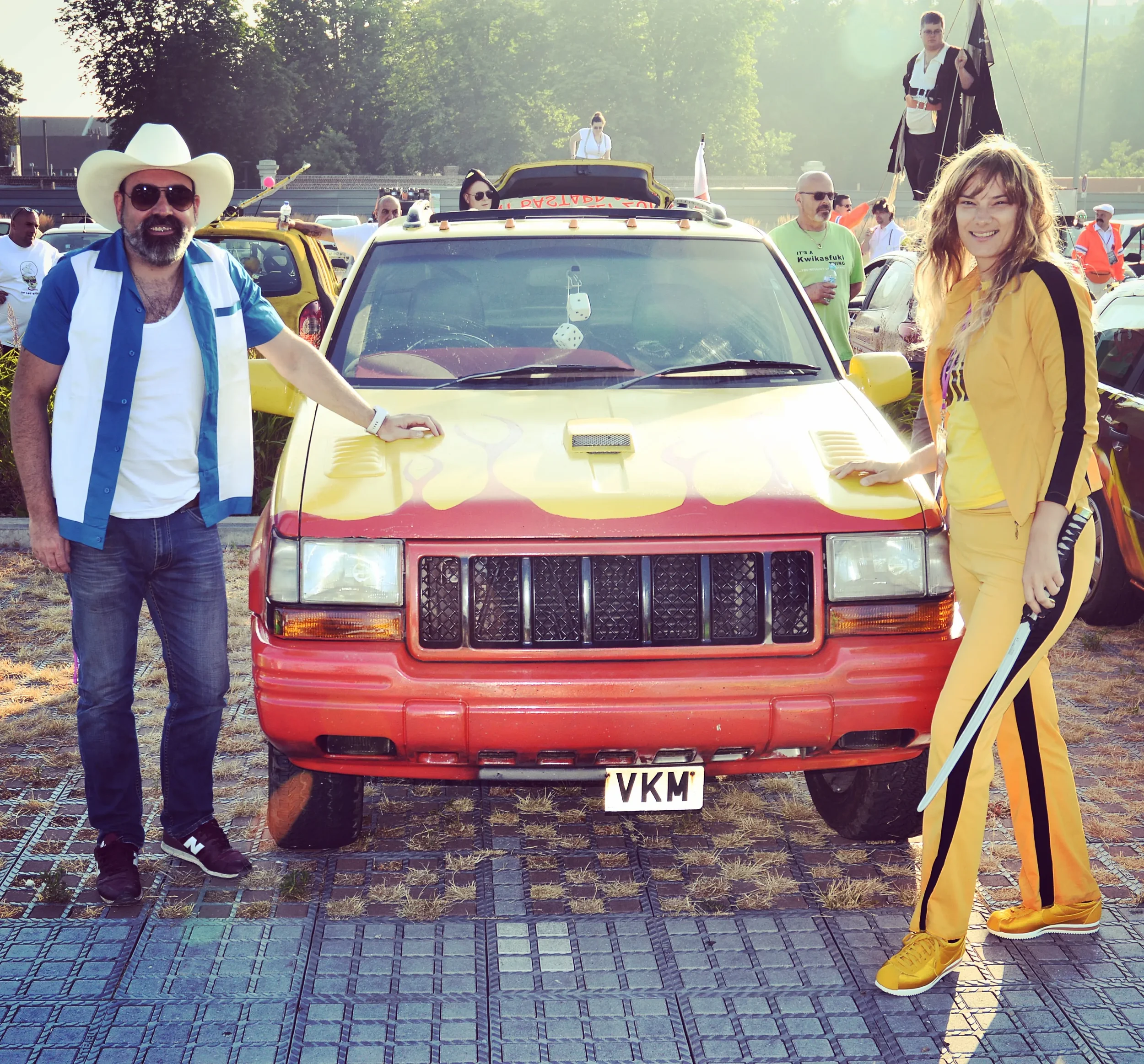 Two people standing on either side of a red and yellow car with a badge reading 'VKM' at a car meet or event. The man on the left is wearing a white cowboy hat, sunglasses, a blue and white shirt, and jeans. The woman on the right is dressed in yello